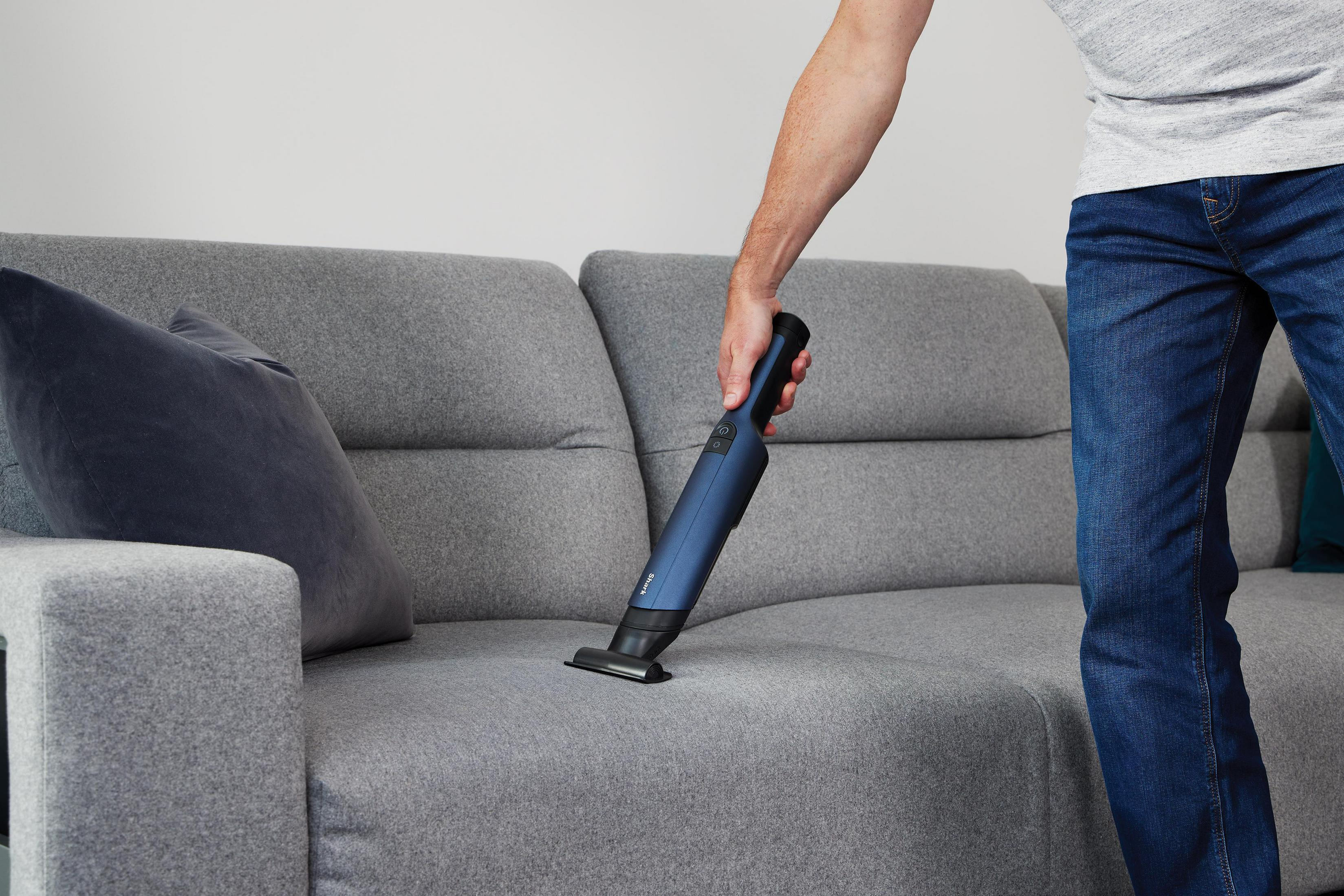 Osoba używająca niebieskiego odkurzacza ręcznego na szarej sofie.