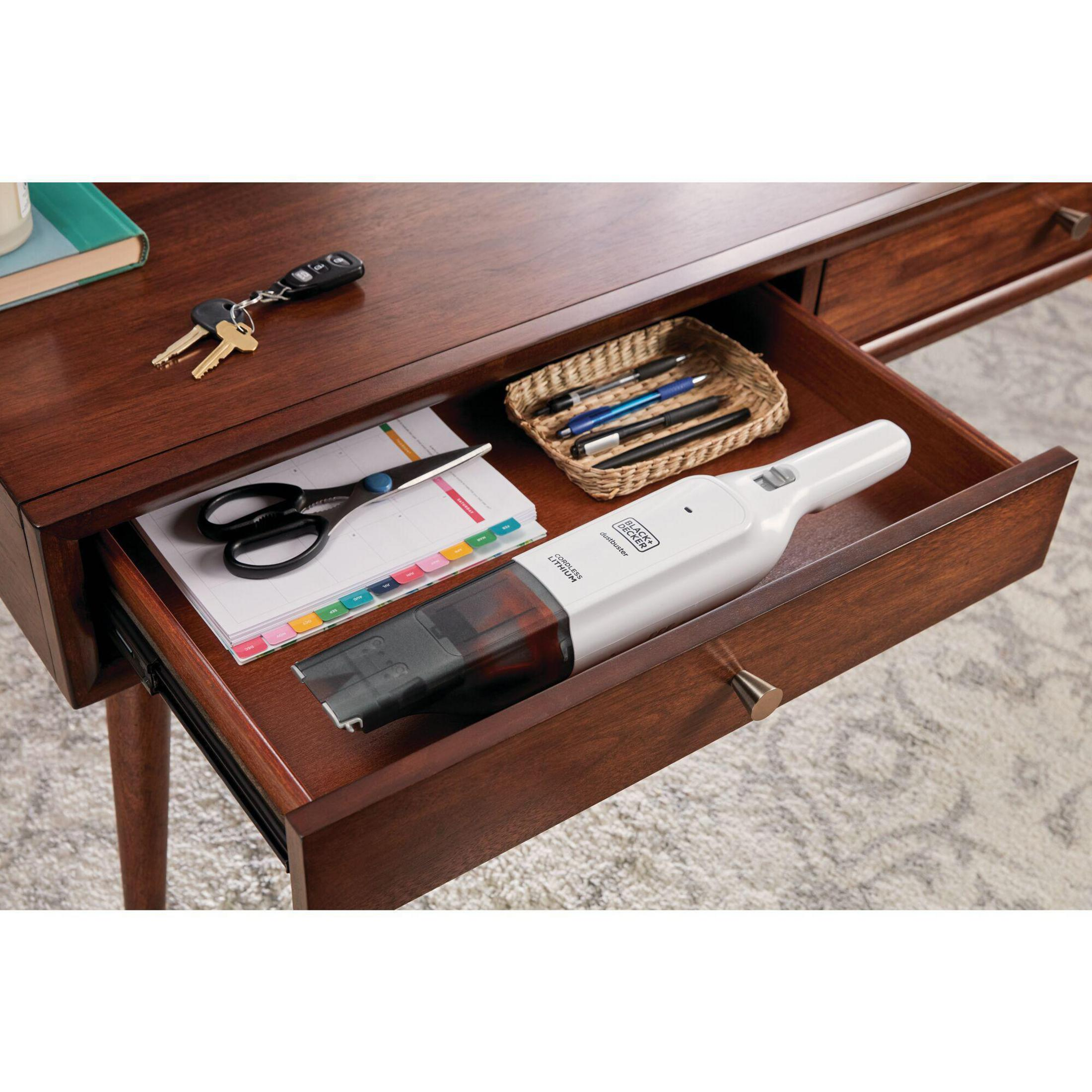 Cassetto aperto con oggetti: forbici, chiavi, penne, aspirapolvere. Scrivania in legno con tappeto grigio sotto.