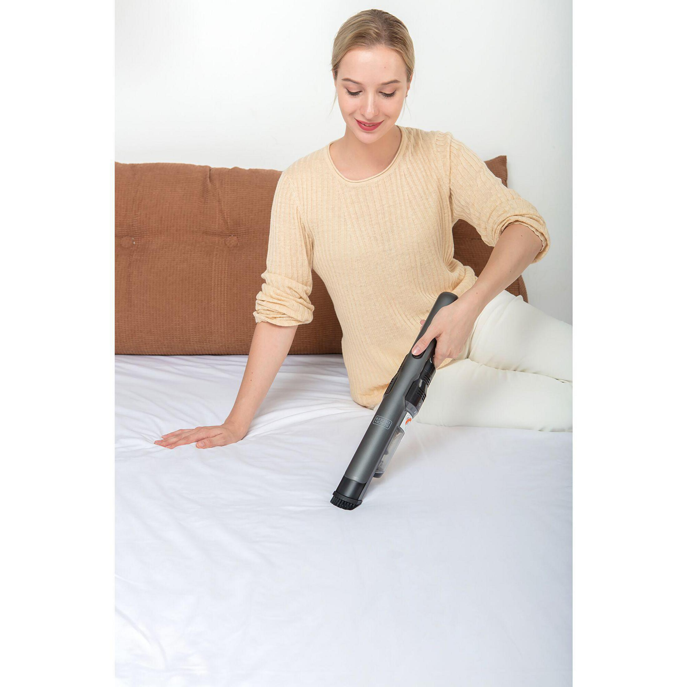 Mujer usando una aspiradora de mano en una cama. Lleva un suéter y pantalones blancos.