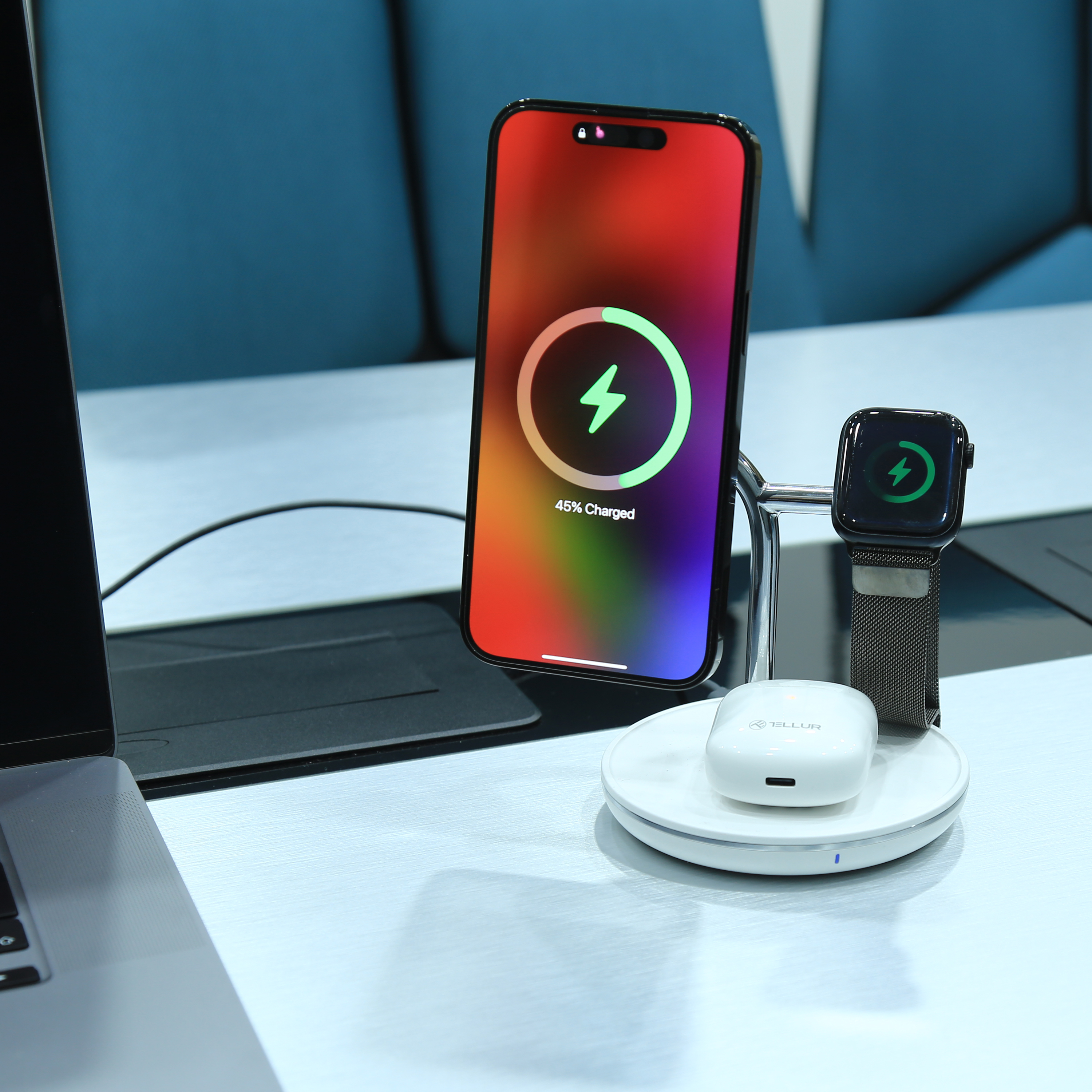 Stacja ładująca z telefonem, zegarkiem i słuchawkami. Telefon pokazuje ikonę ładowania. Biała powierzchnia, niebieskie tło.