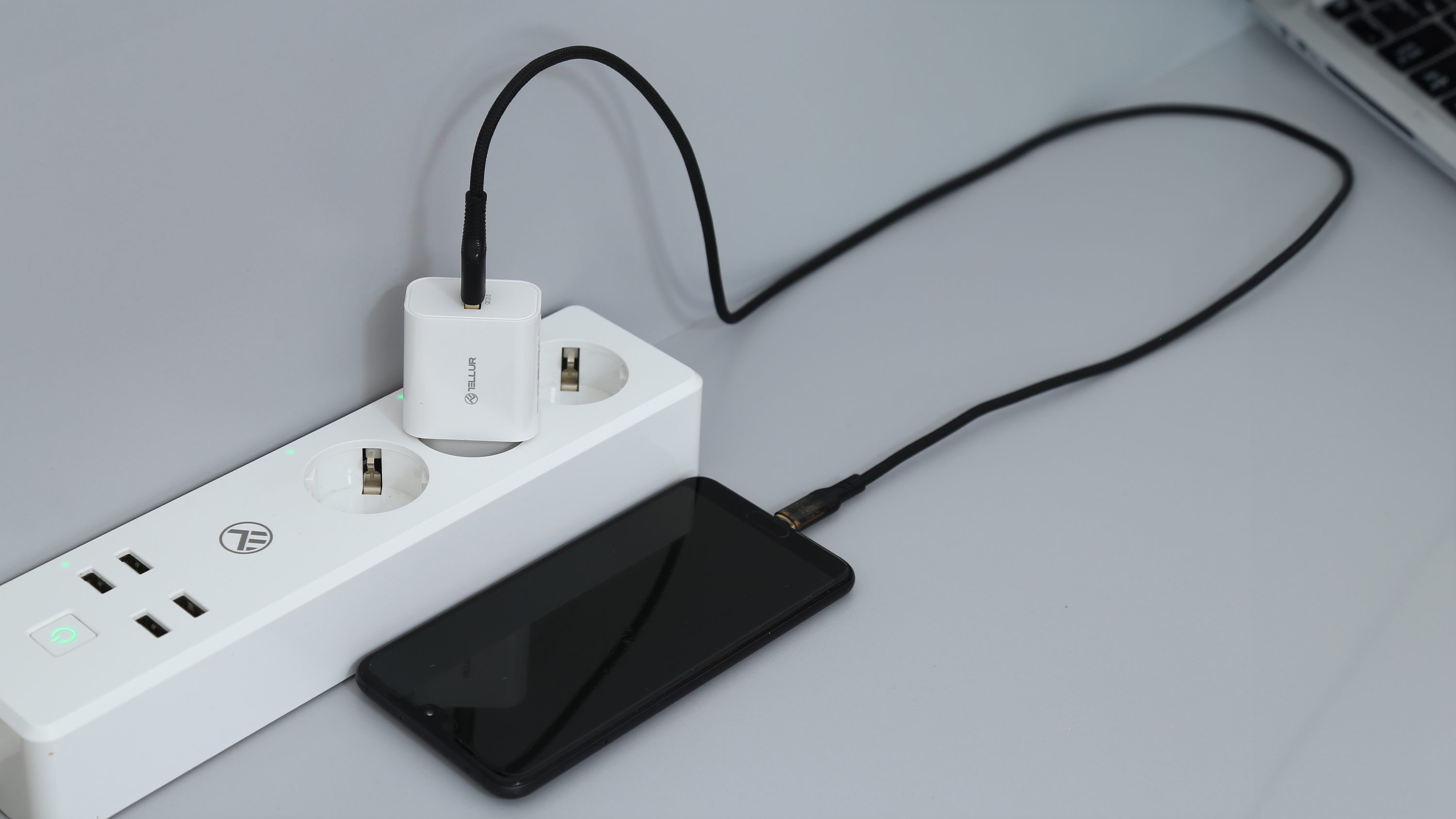 Biała listwa zasilająca z telefonem ładującym się za pomocą czarnego kabla i białej ładowarki.