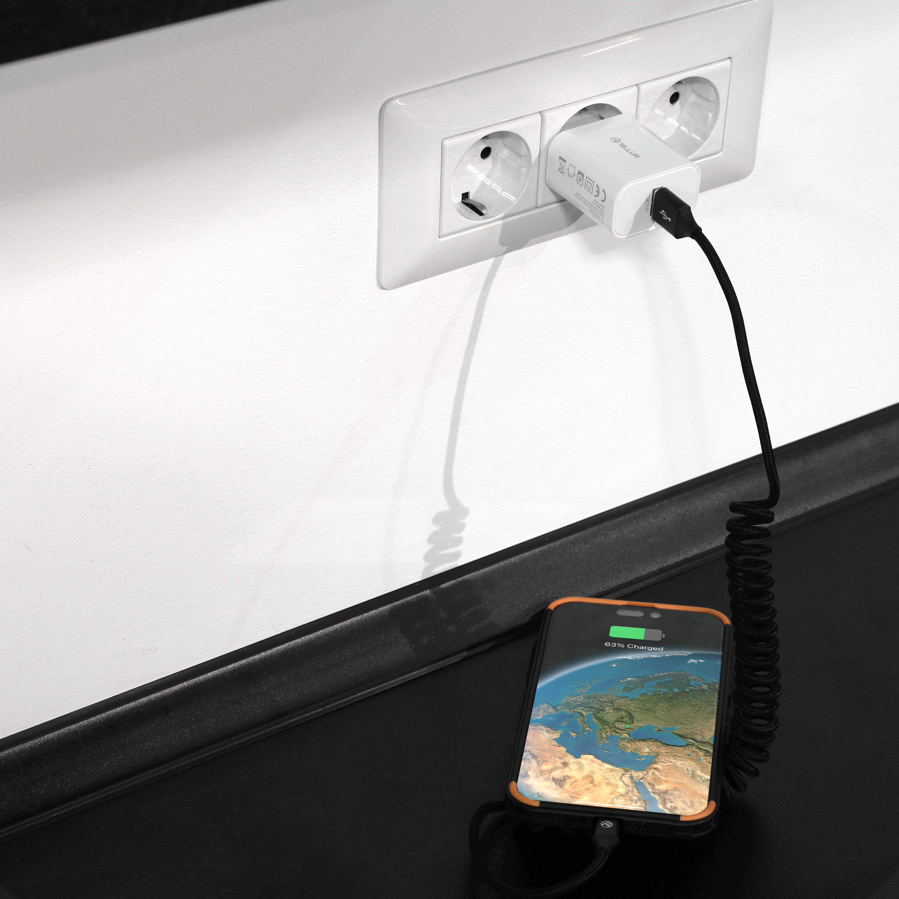 Telefon ładujący się czarnym kablem, podłączony do białego gniazdka na ścianie.