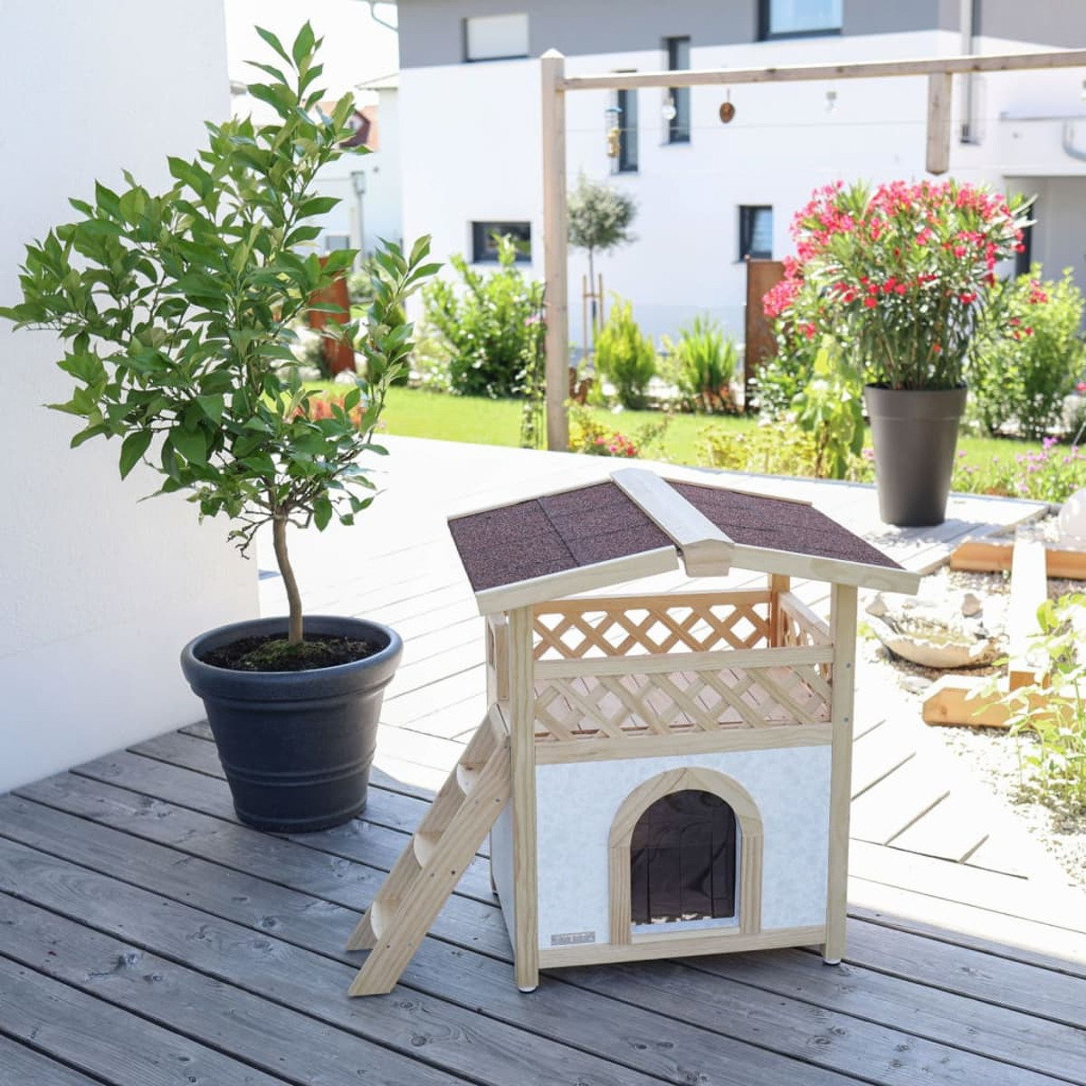 Holzhundehütte mit Leiter neben einer Topfpflanze auf einer Holzterrasse. Im Hintergrund ein Haus und Bäume.