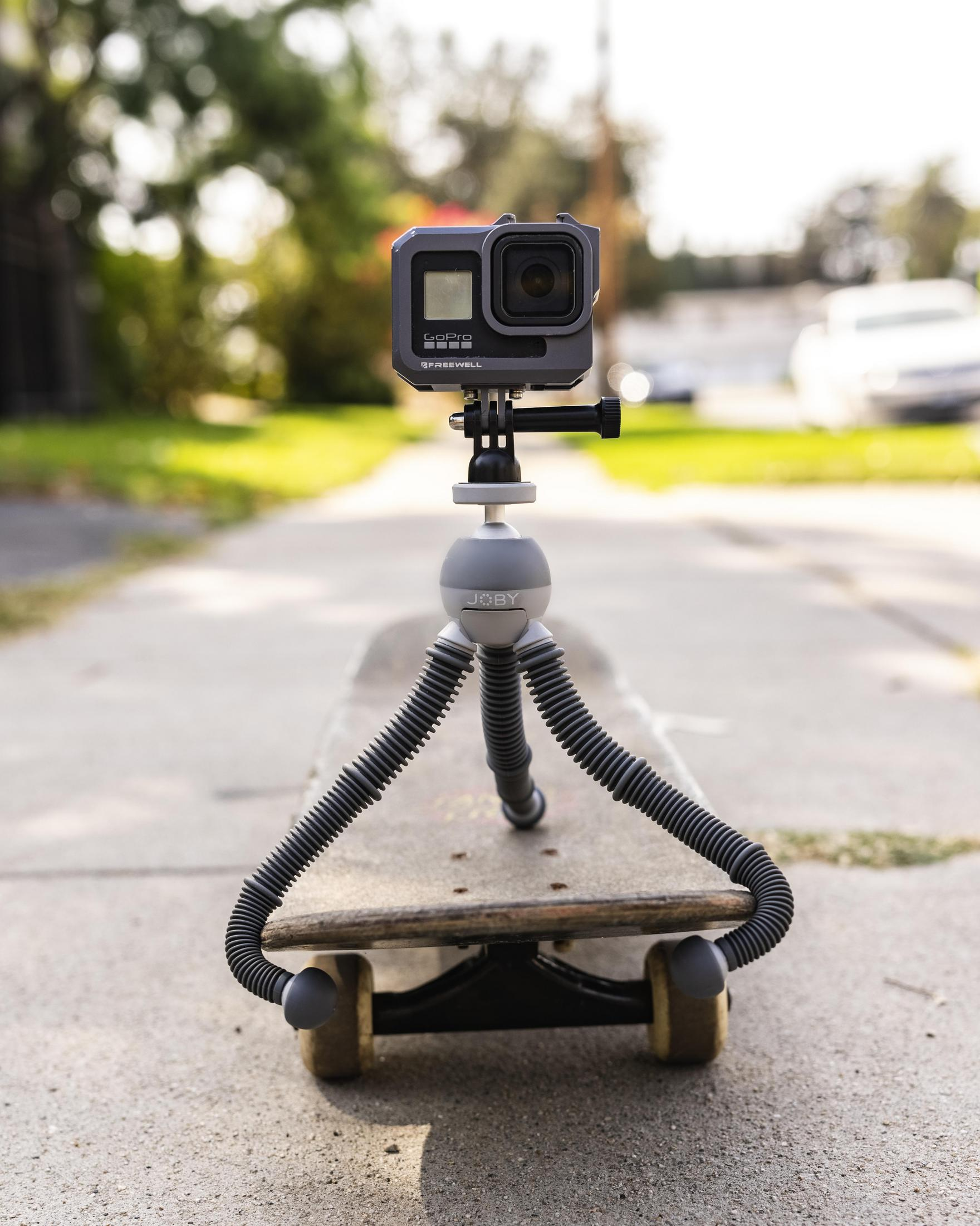 Caméra GoPro sur un trépied monté sur un skateboard sur un trottoir. Arrière-plan: arbres et une route.