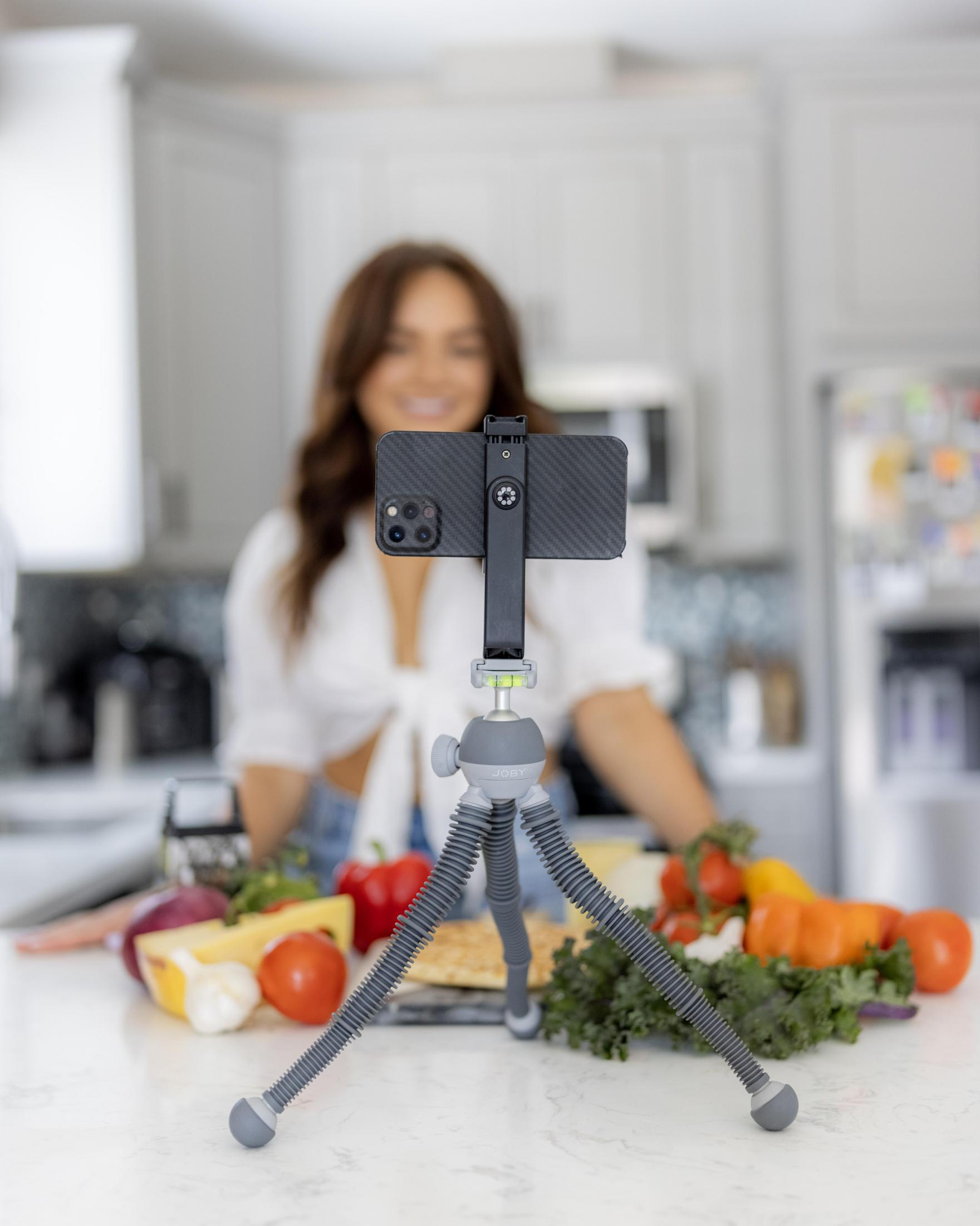 Kobieta filmuje w kuchni z statywem, telefonem. Warzywa, ser na stole. Rozmyte tło.