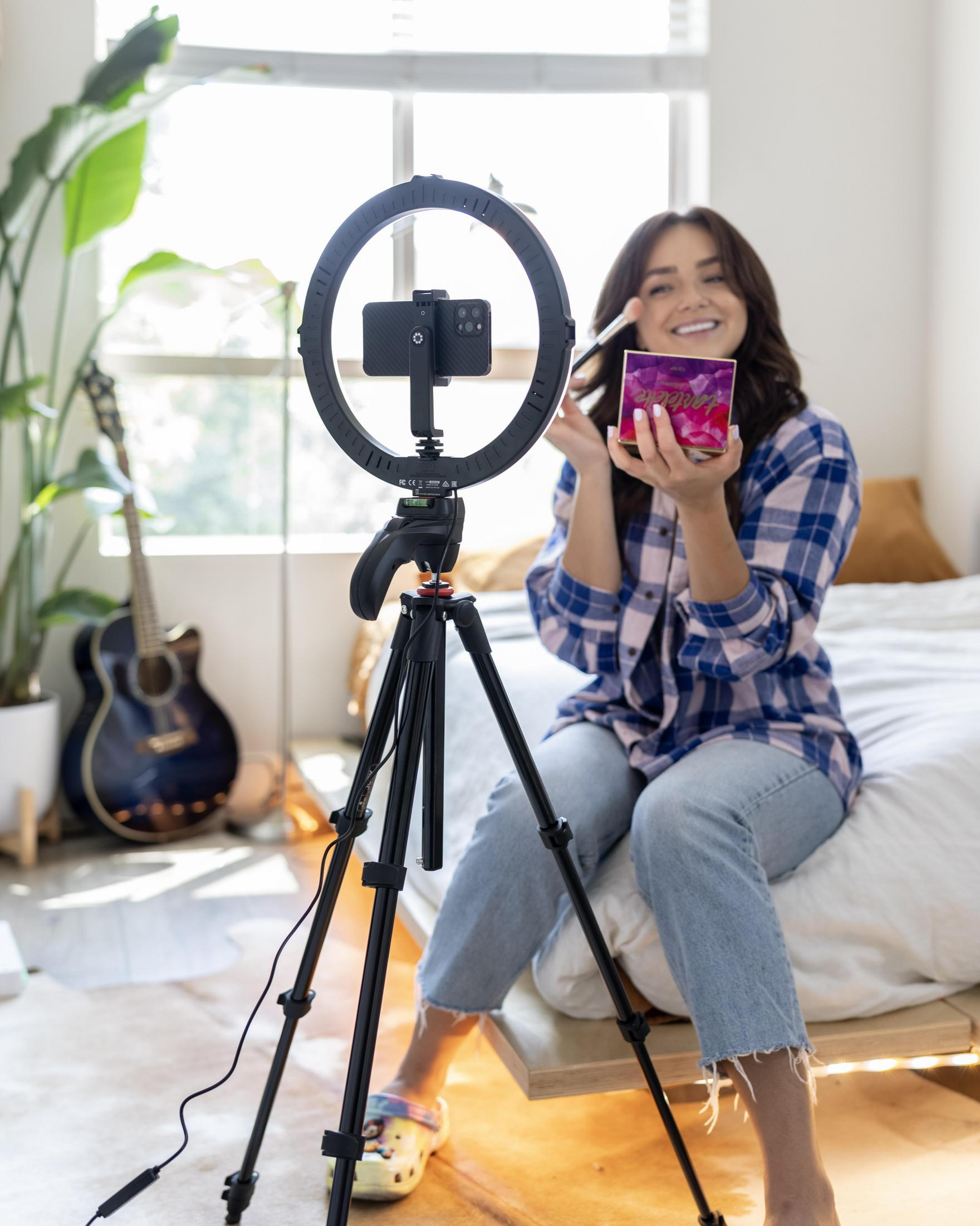 Kobieta z ringiem, telefonem i pędzlem do makijażu. Gitara i łóżko w tle. Jasne okno.