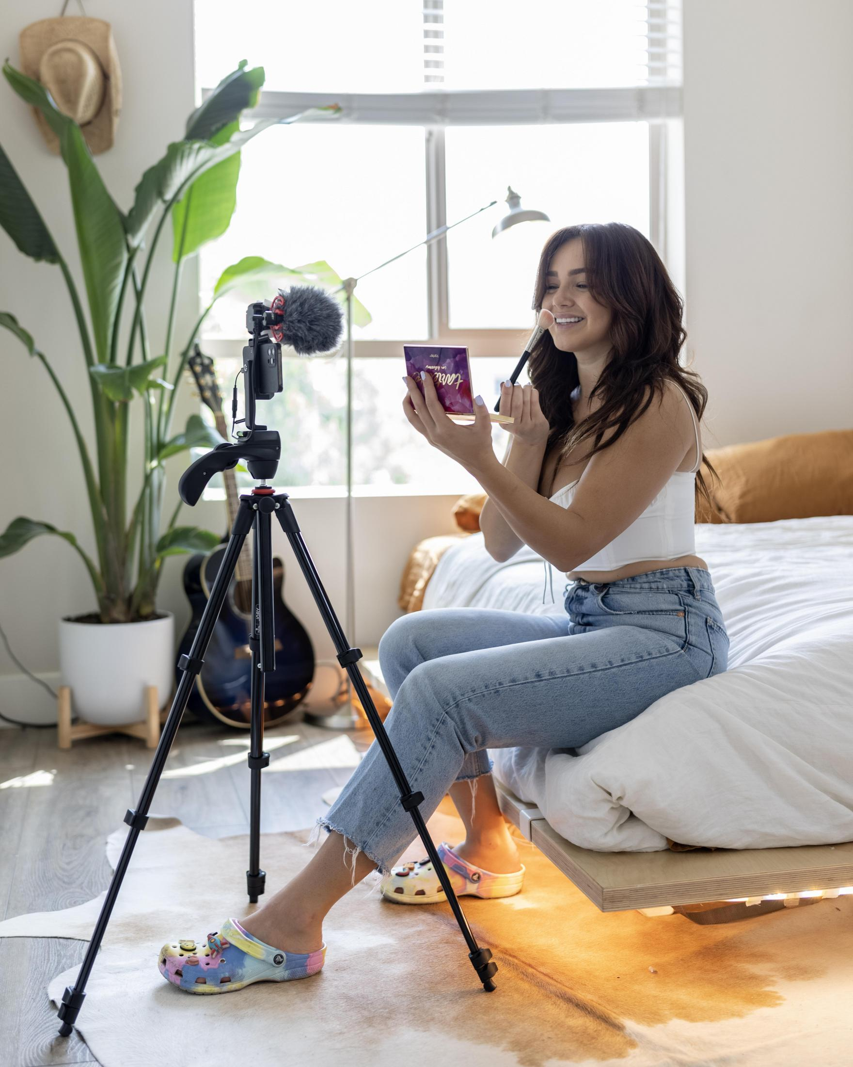 Kobieta nakładająca makijaż podczas nagrywania wideobloga, z kamerą i mikrofonem. Tło obejmuje łóżko i rośliny.