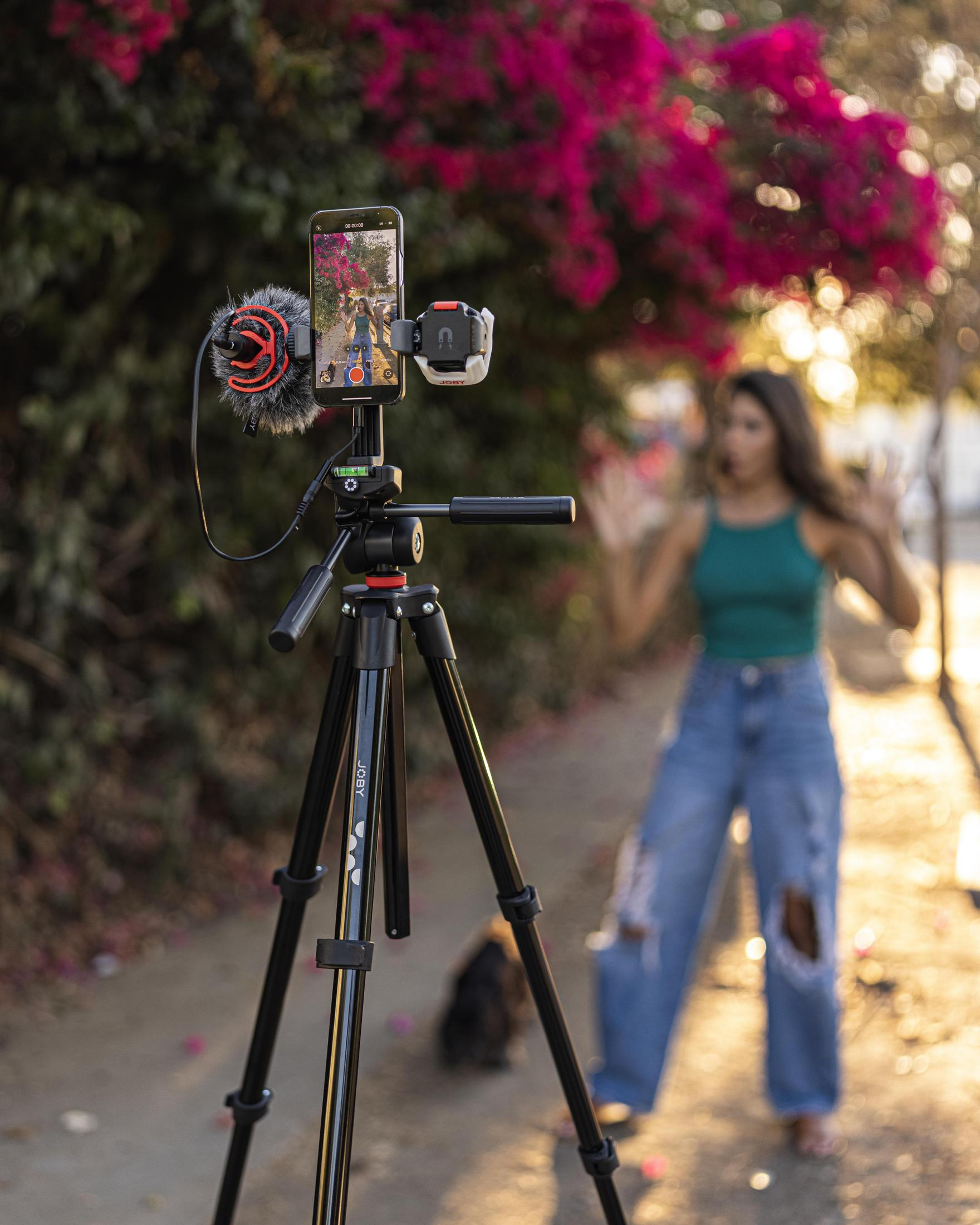 Vlogger nagrywa telefonem na statywie. Kobieta jest niewyraźna w tle.