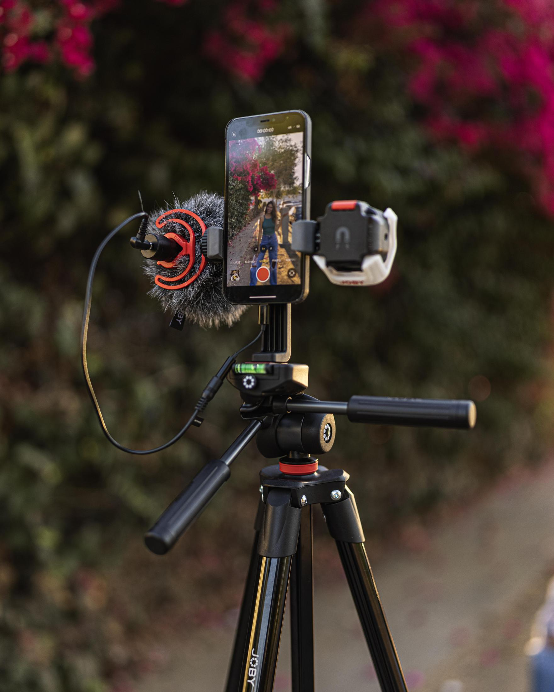 Telefon nagrywający kobietę na zewnątrz, zamontowany na statywie z mikrofonem. Tło z rozmytymi liśćmi.