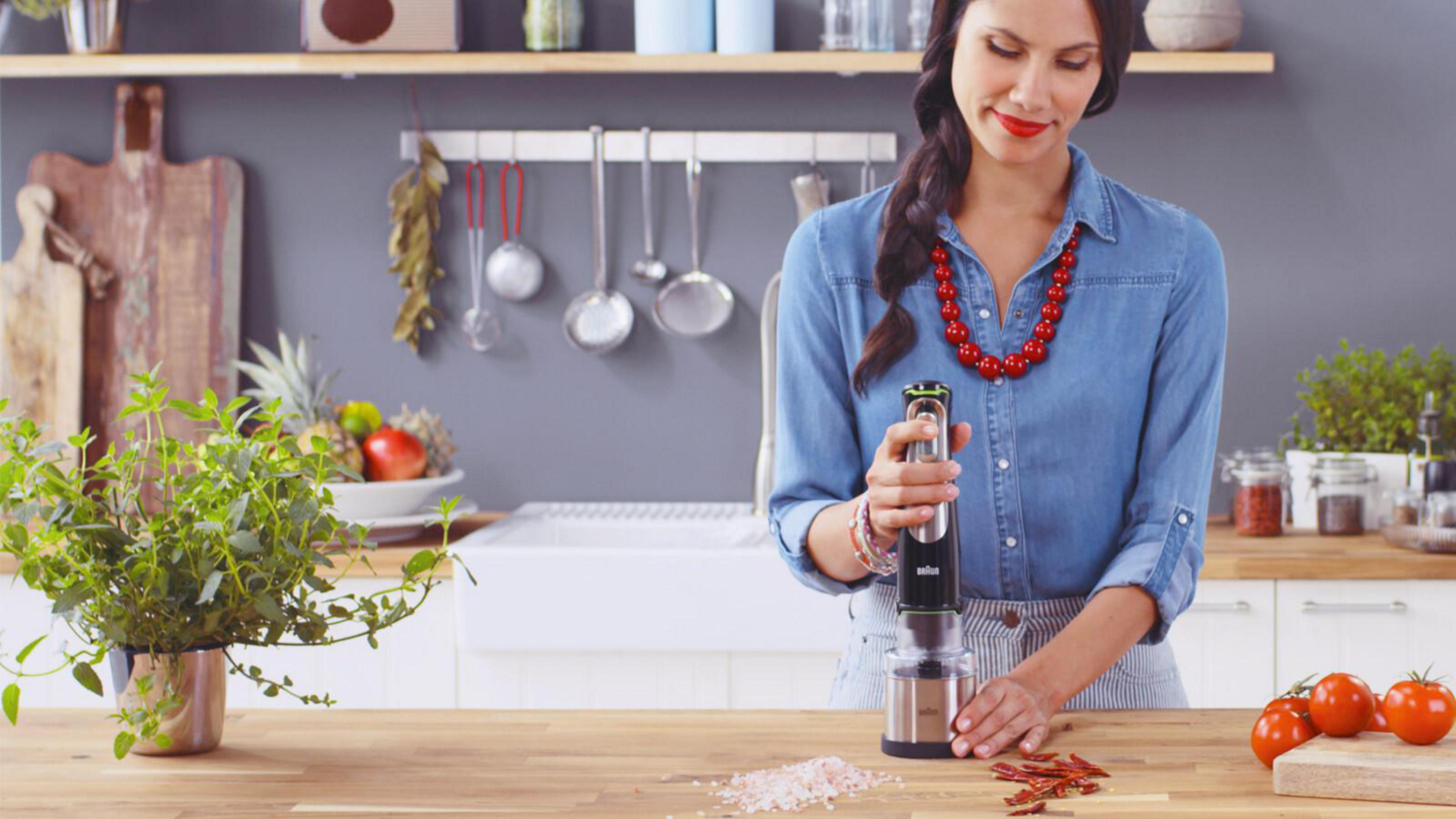 Donna che macina spezie con un frullatore a immersione Braun. Collana rossa, pomodori ed erbe visibili.