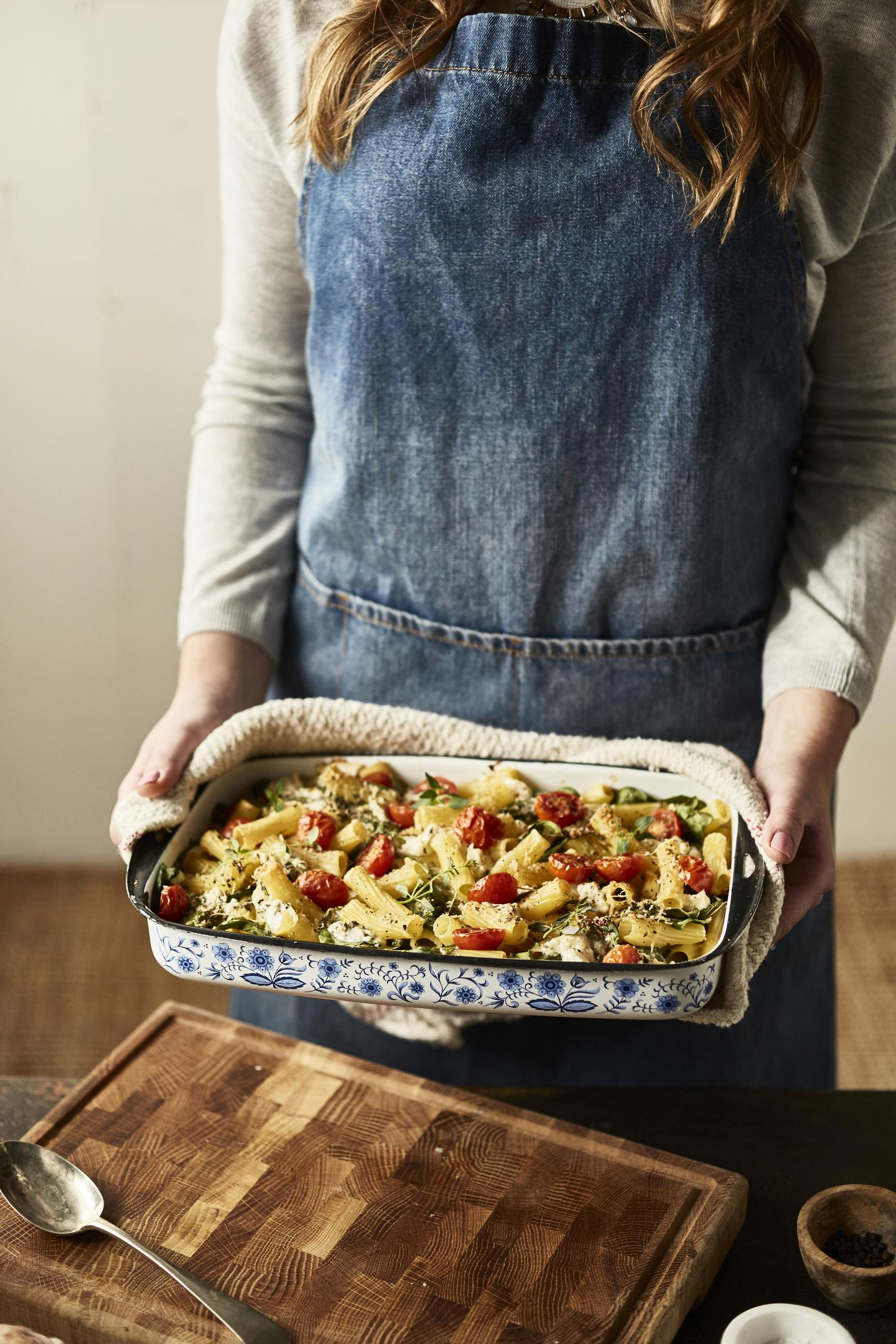 Osoba trzymająca naczynie żaroodporne z makaronem, pomidorami i ziołami.