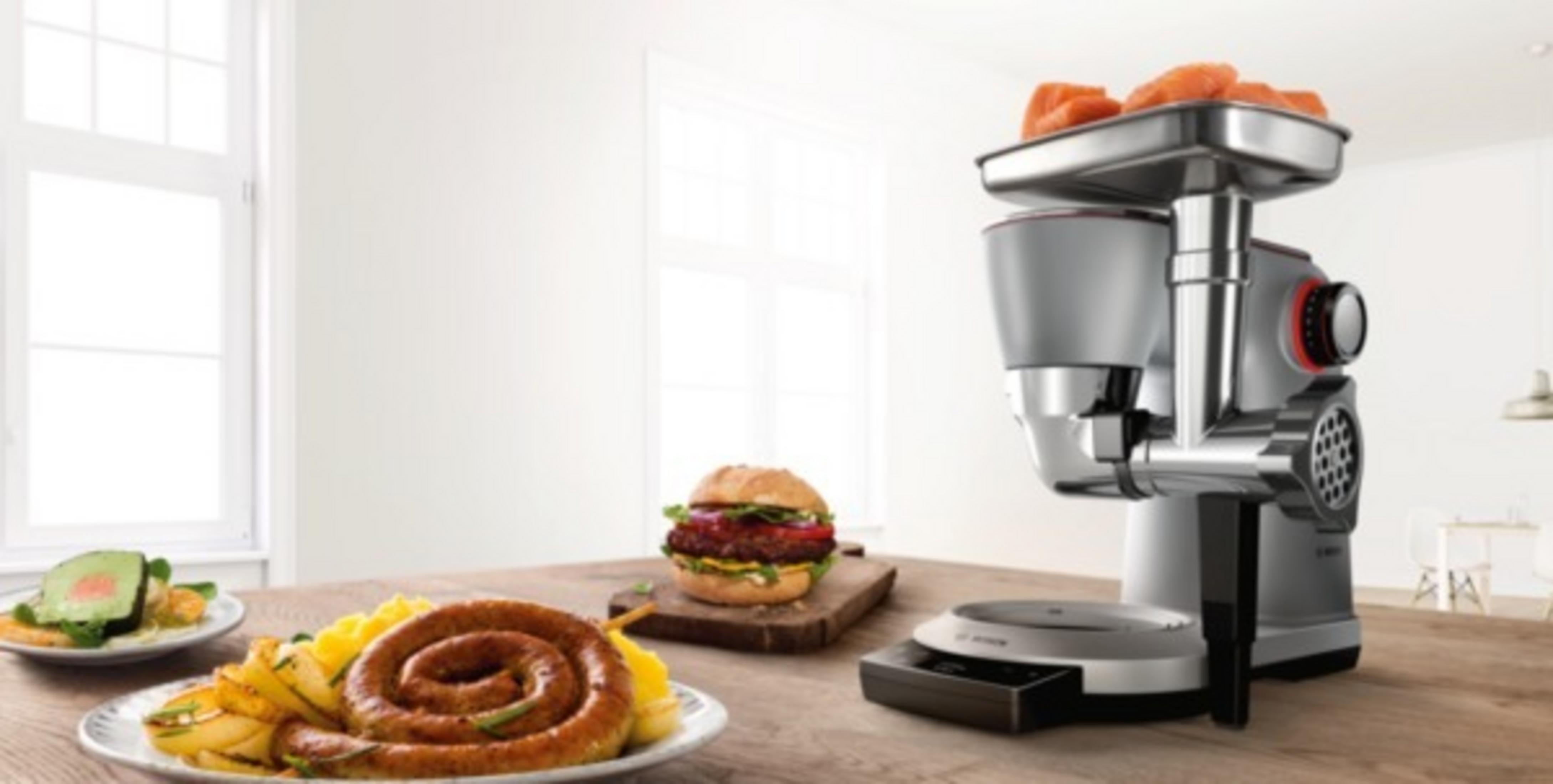 Küchenmaschine mit Wurst, Burger und Zutaten auf Holztisch. Weiße Theke sichtbar.