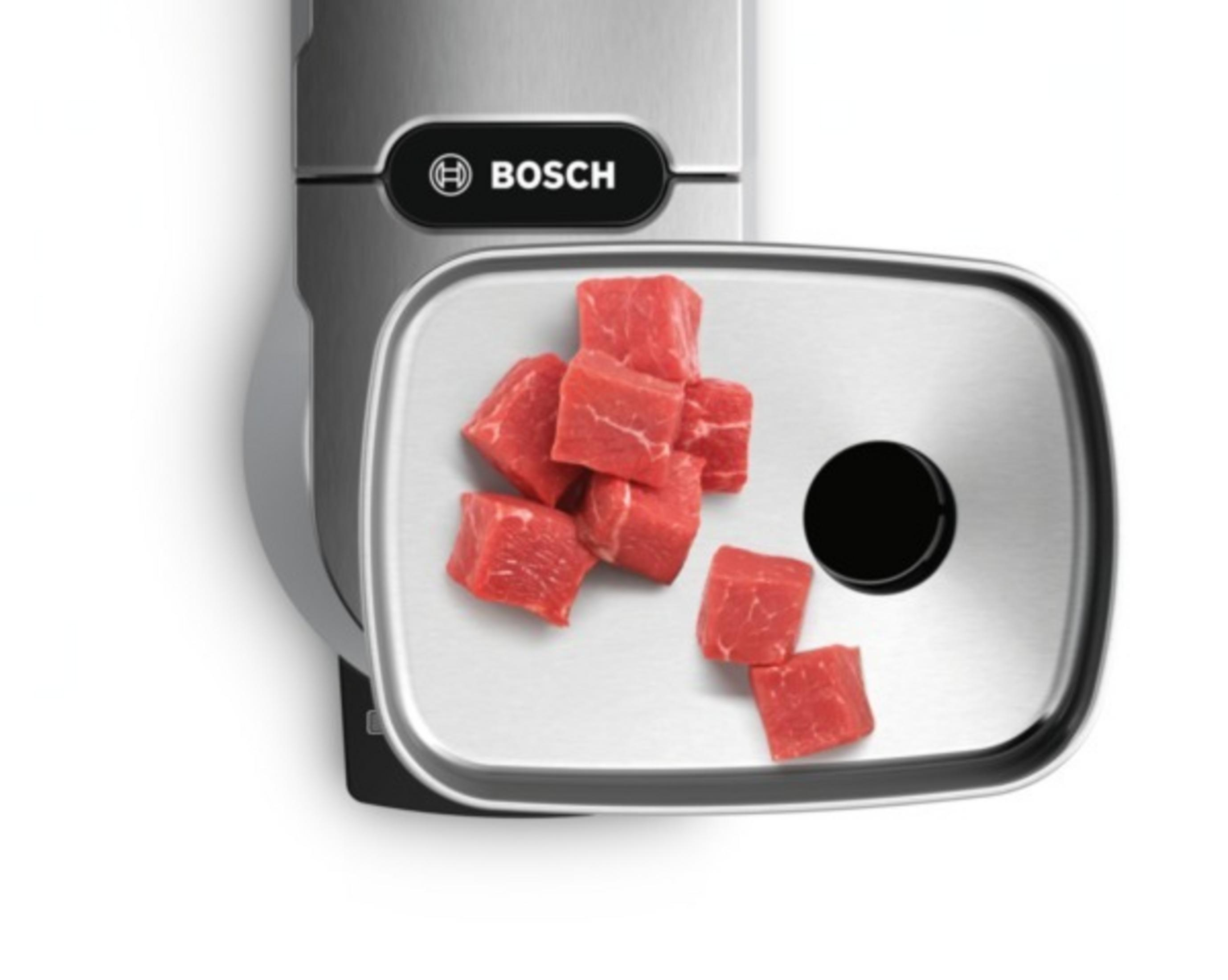 Metallschale eines Fleischwolfs mit rohen Fleischwürfeln neben der Bosch Maschine.