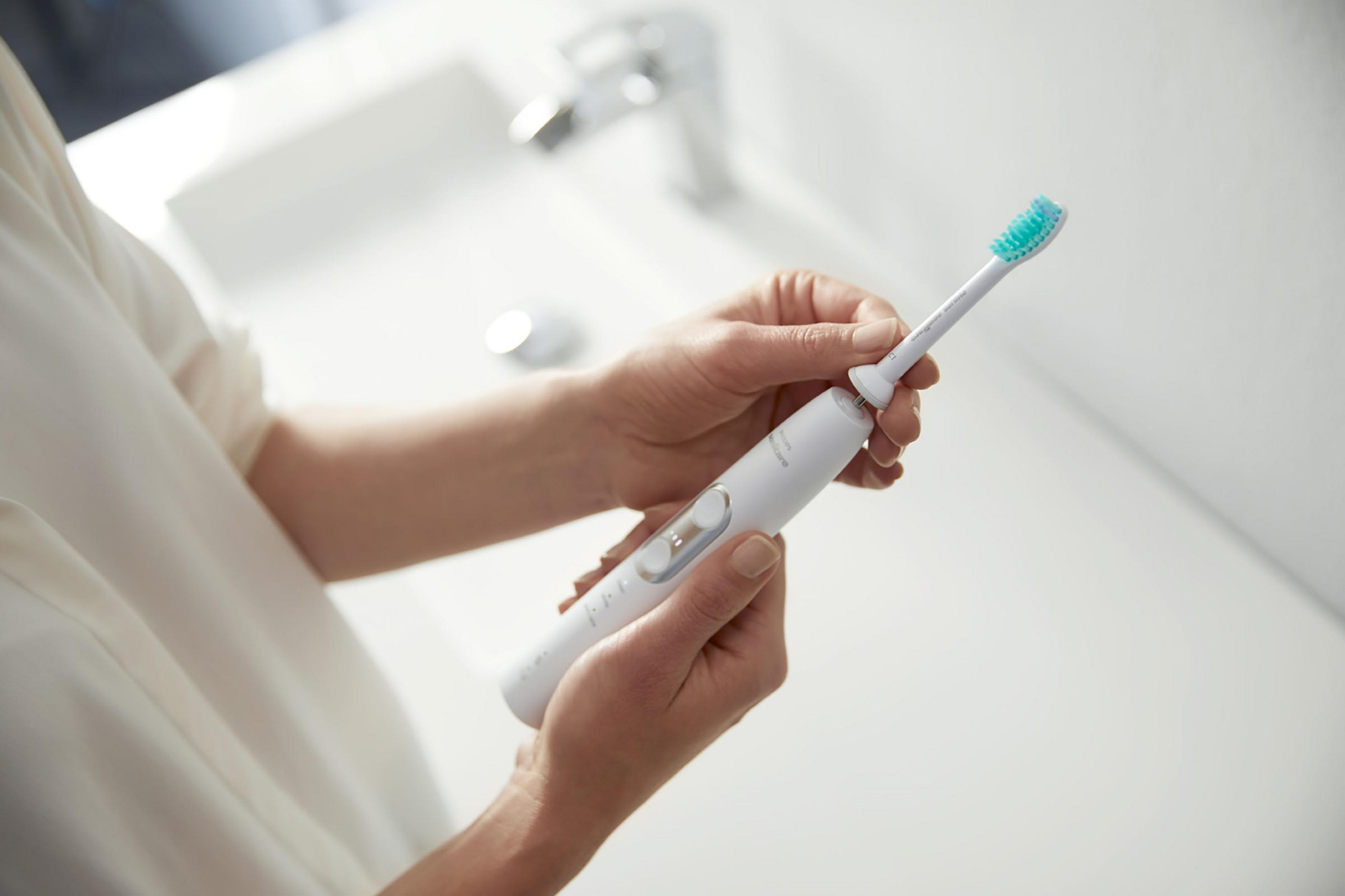 Una persona sostiene un cepillo de dientes eléctrico blanco y plateado sobre un lavabo blanco.