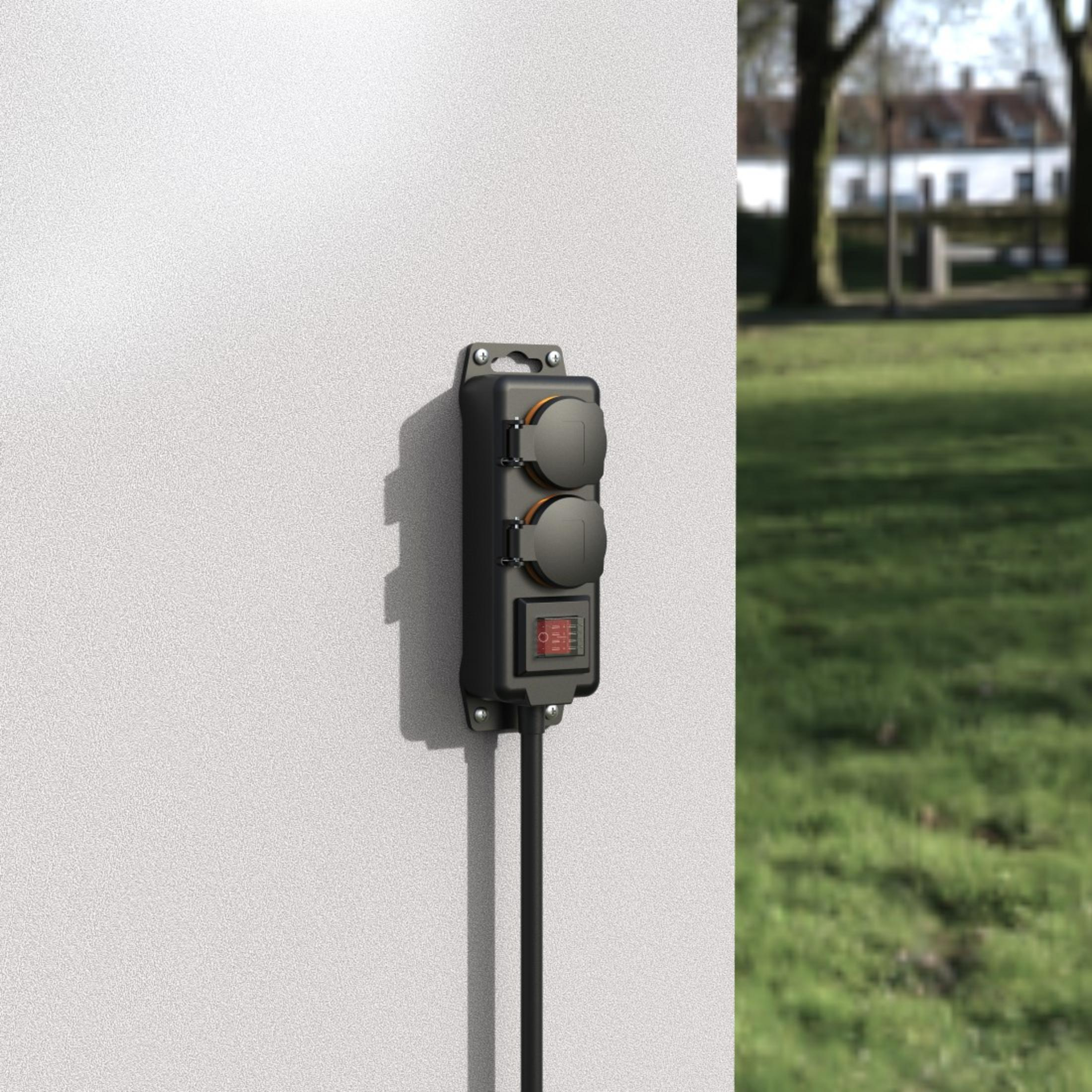 Czarna skrzynka gniazdka elektrycznego zamontowana na białej ścianie. Widoczny czerwony przełącznik. Zielona trawa w tle.