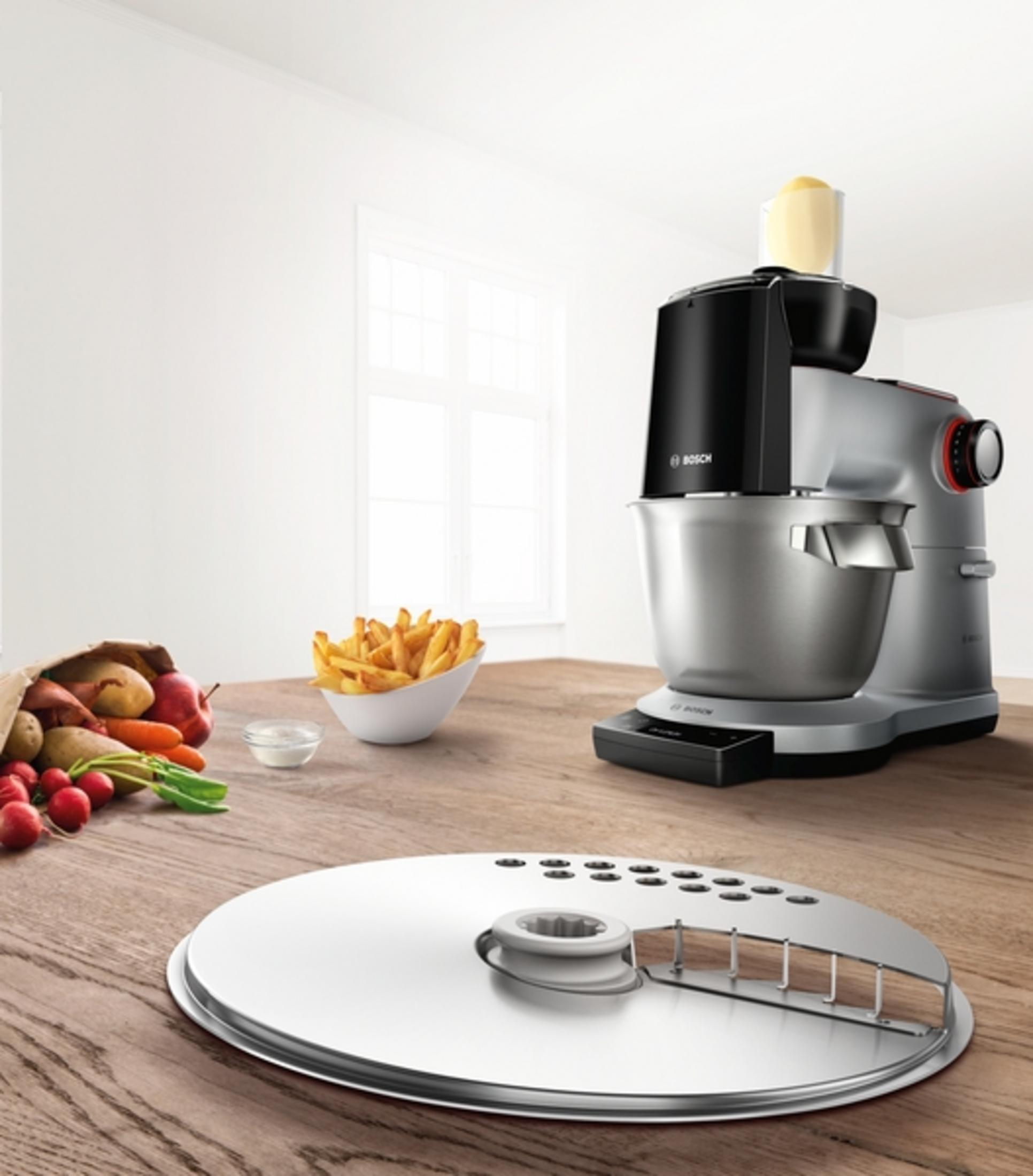Bosch Küchenmaschine mit Gemüse, Pommes und einem Aufsatz auf einem Holztisch.