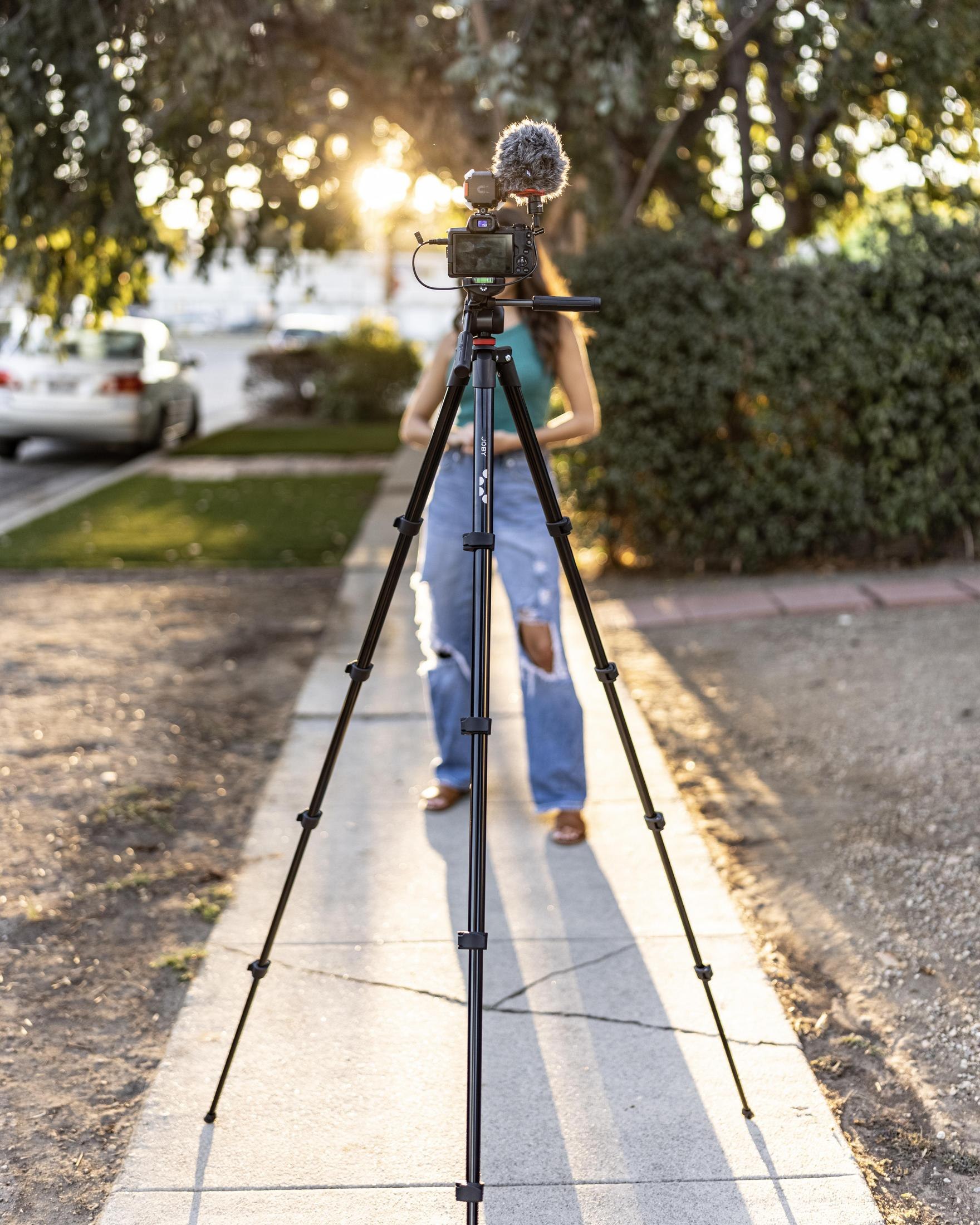 Mujer filmando afuera con cámara en trípode. Sol brillante en el fondo, entorno callejero.