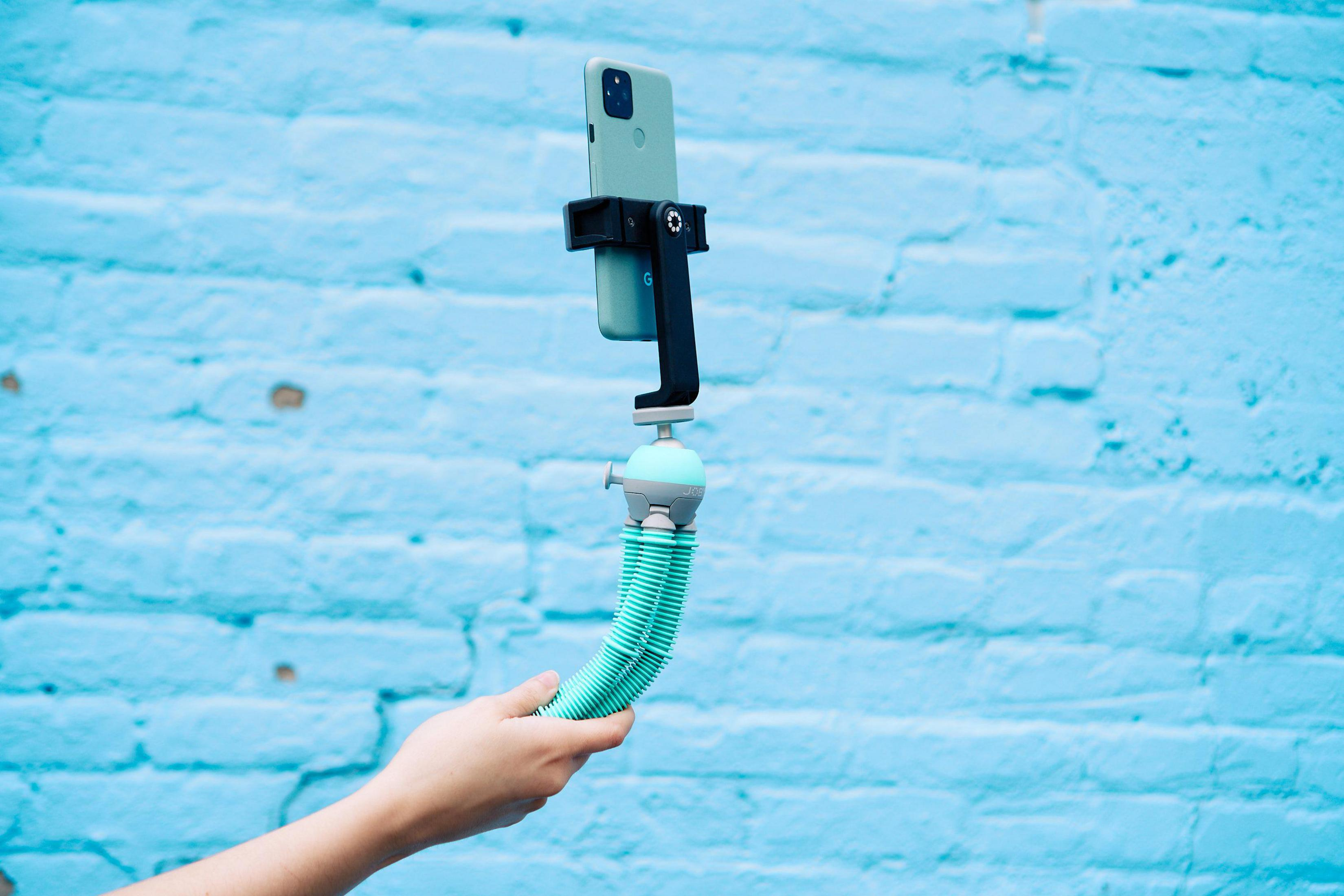 Person hält Telefon auf flexiblem Stativ. Blauer Backsteinmauer-Hintergrund.