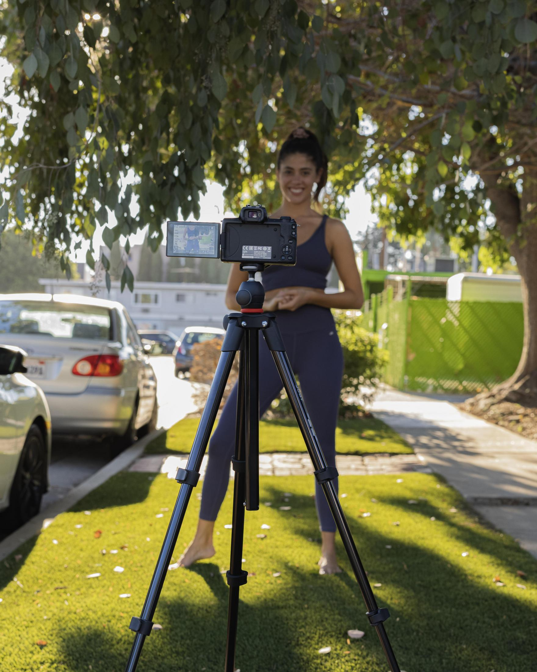 Kobieta filmująca siebie kamerą na statywie w podmiejskim otoczeniu.