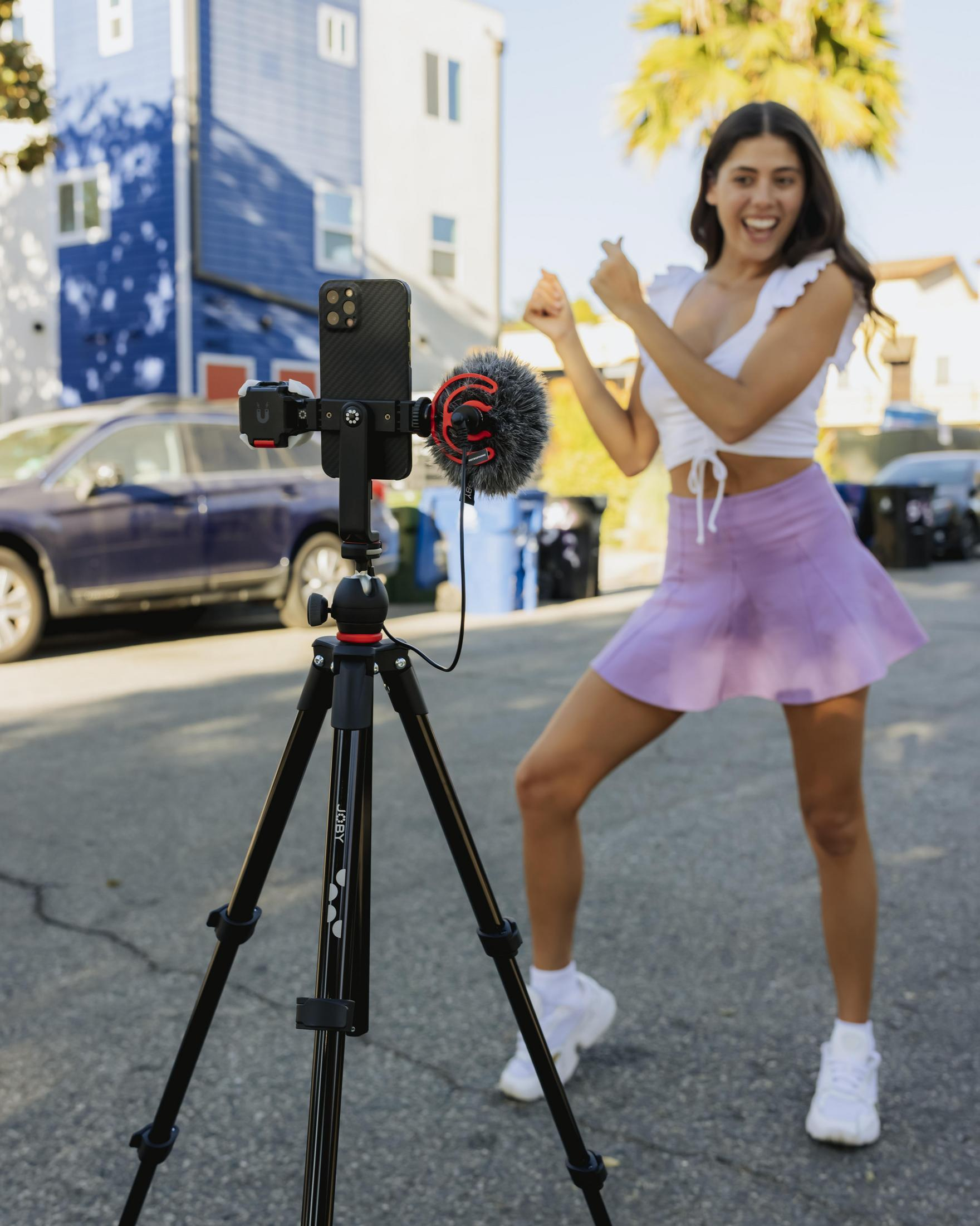 Kobieta tańczy, filmując telefonem na statywie. Tło: ulica miejska.