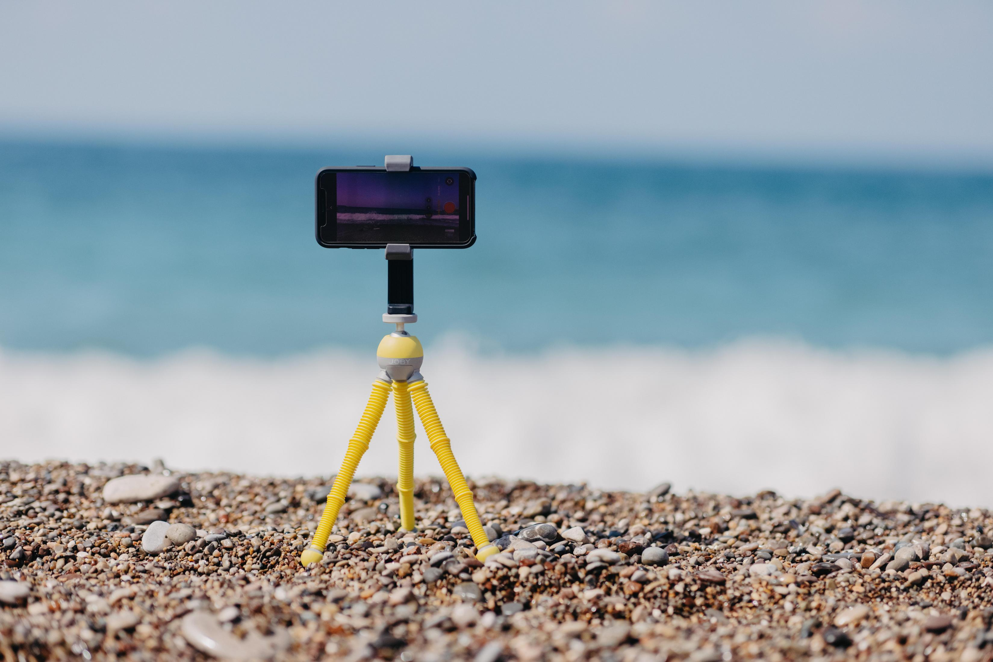 Smartphone en un trípode amarillo en la playa, con fondo de océano azul.