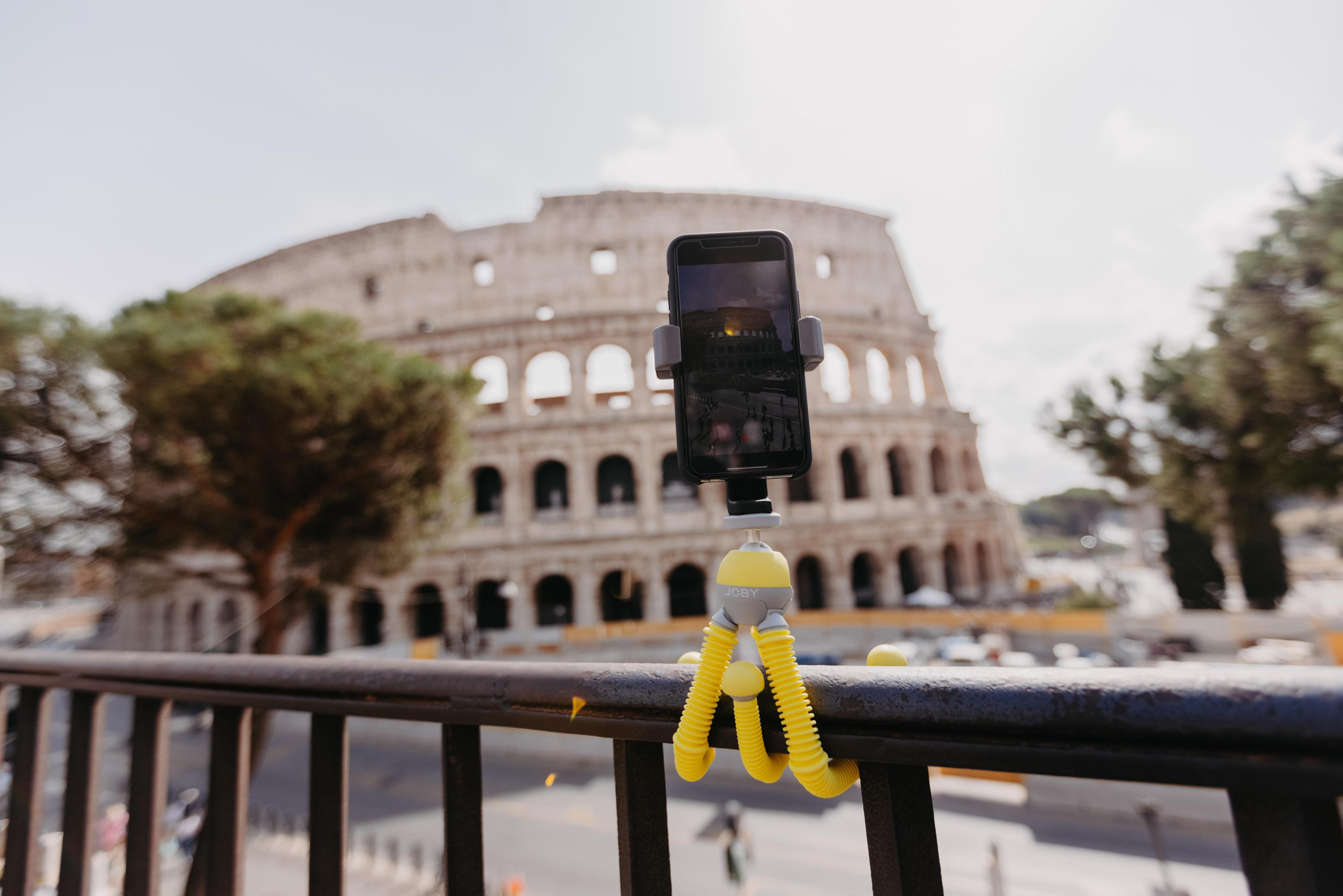 Teléfono en trípode amarillo, apoyado en una barandilla. Coliseo al fondo.