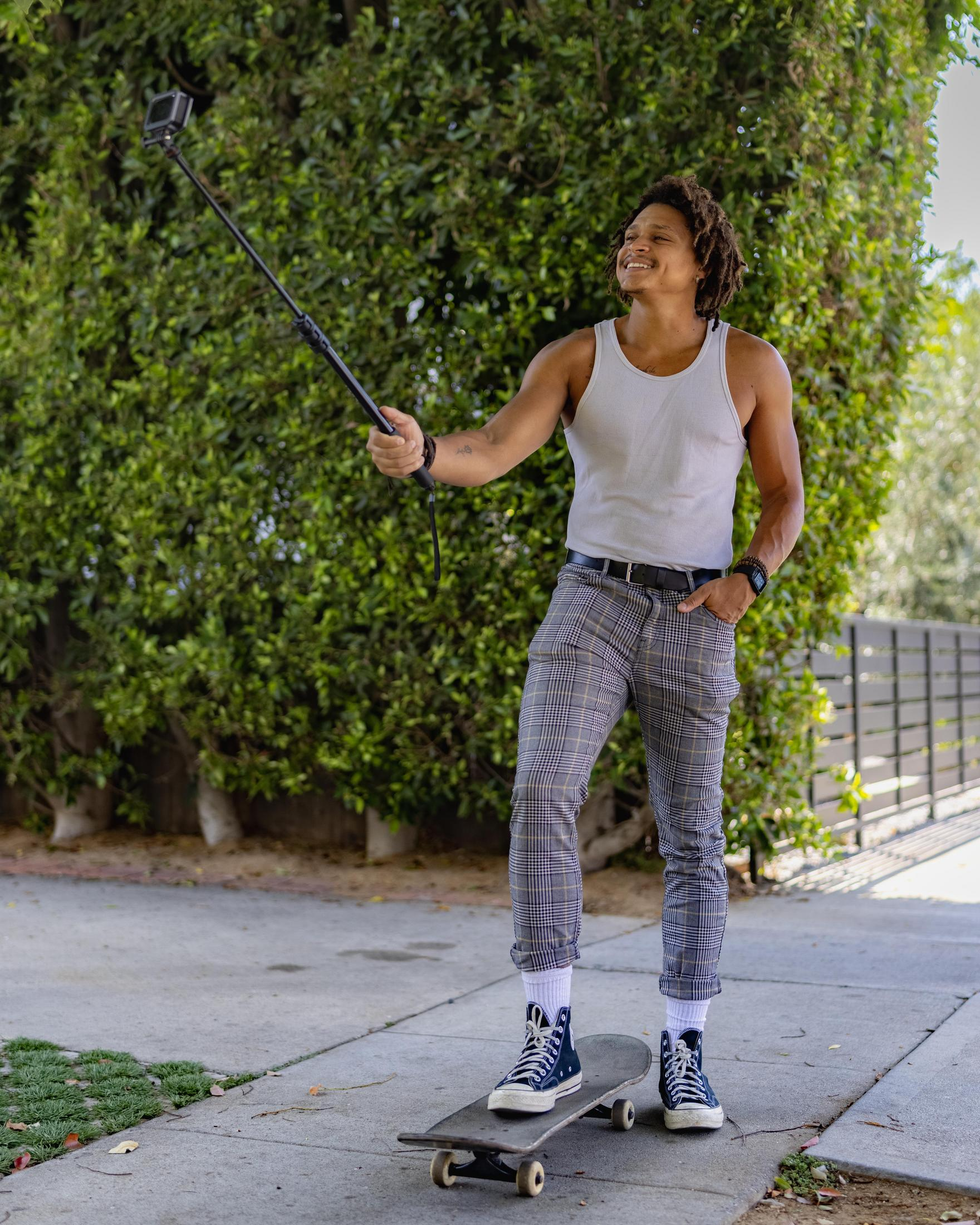 Man on skateboard holding a selfie stick, smiling. Green bushes in background.