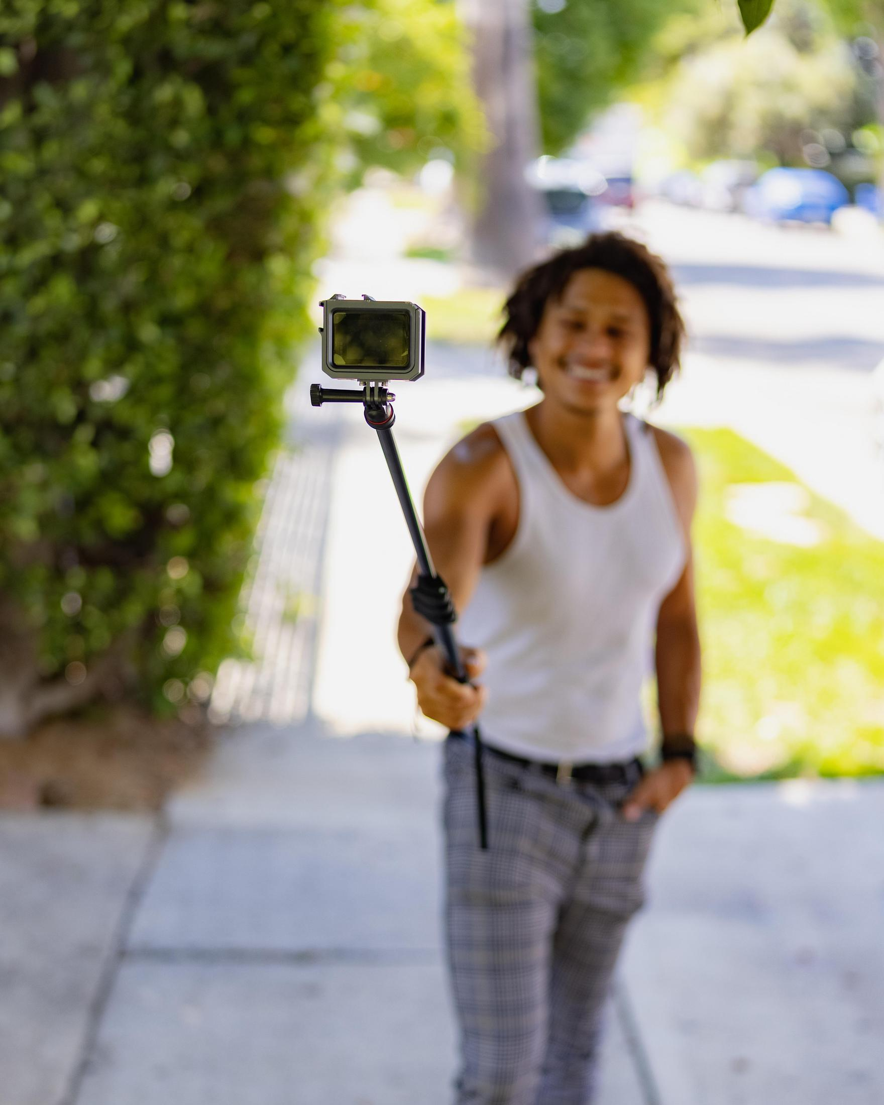Man holding a selfie stick with a camera, smiling. The background is blurry.
