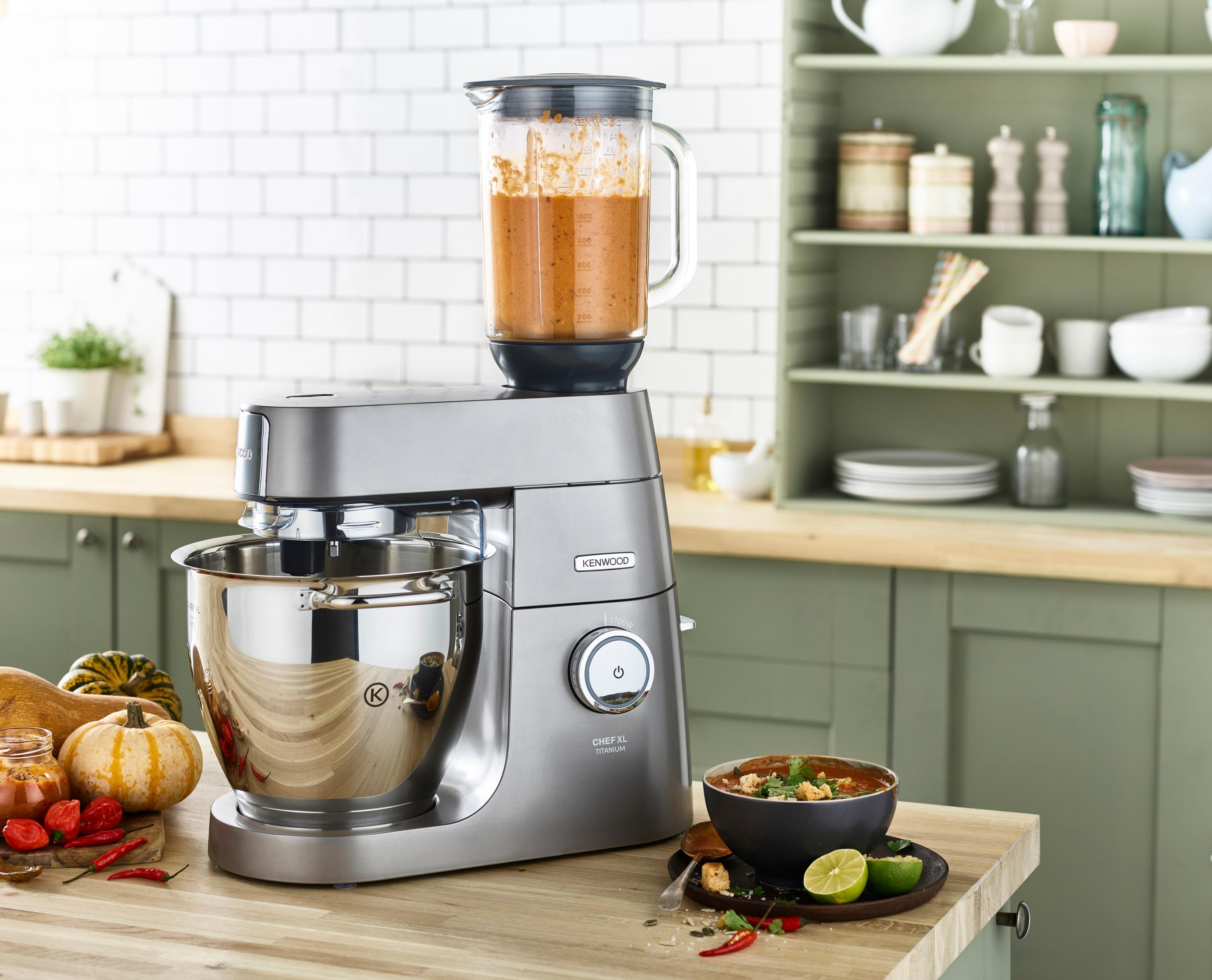 Silberner Kenwood-Mixer mit Blender oben. Küchenumgebung mit Zutaten. Holztisch. Grüne Schränke.