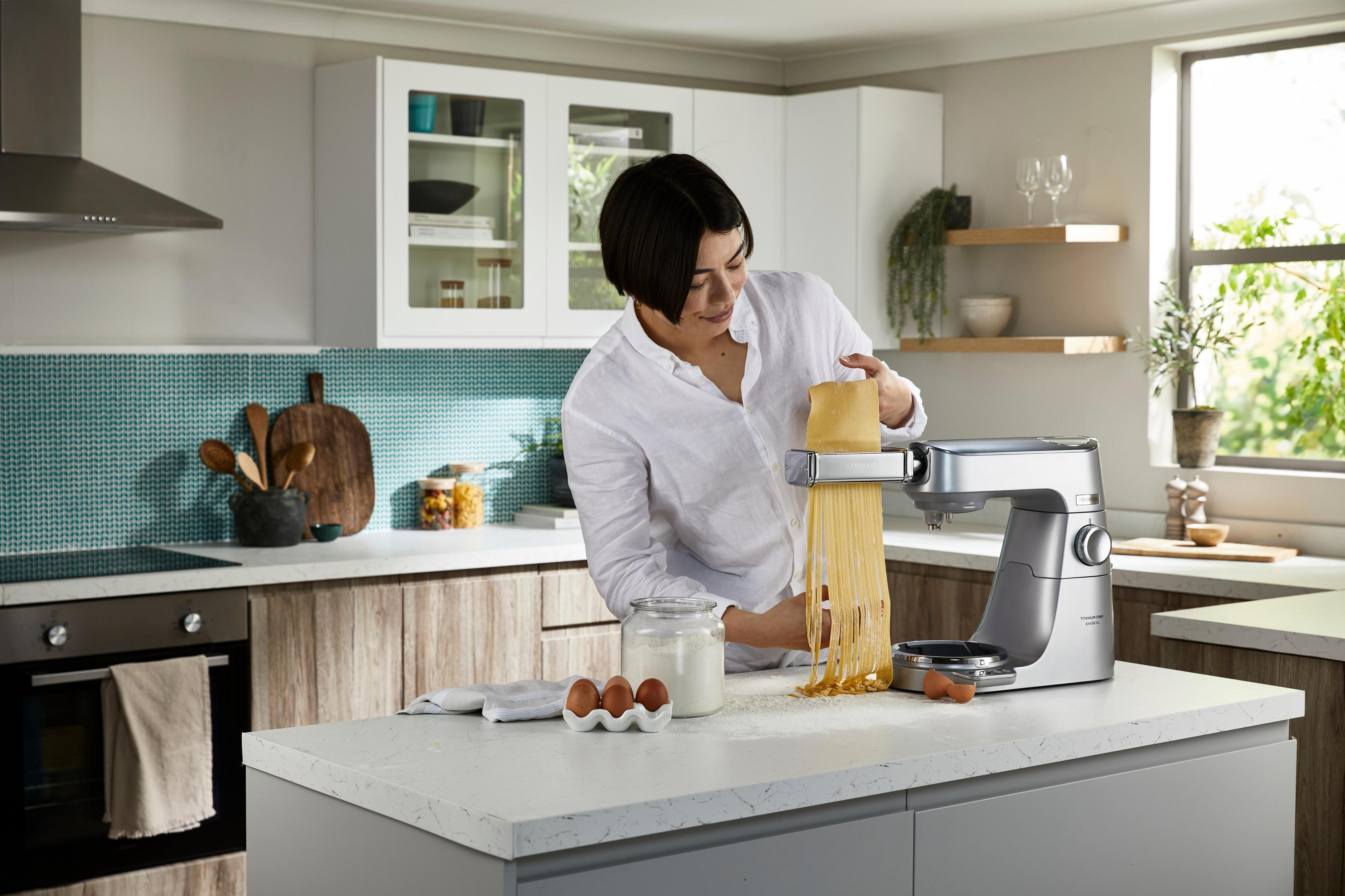 Kobieta używa maszynki do makaronu w kuchni. Składniki, szafki i sprzęty są widoczne w tle.
