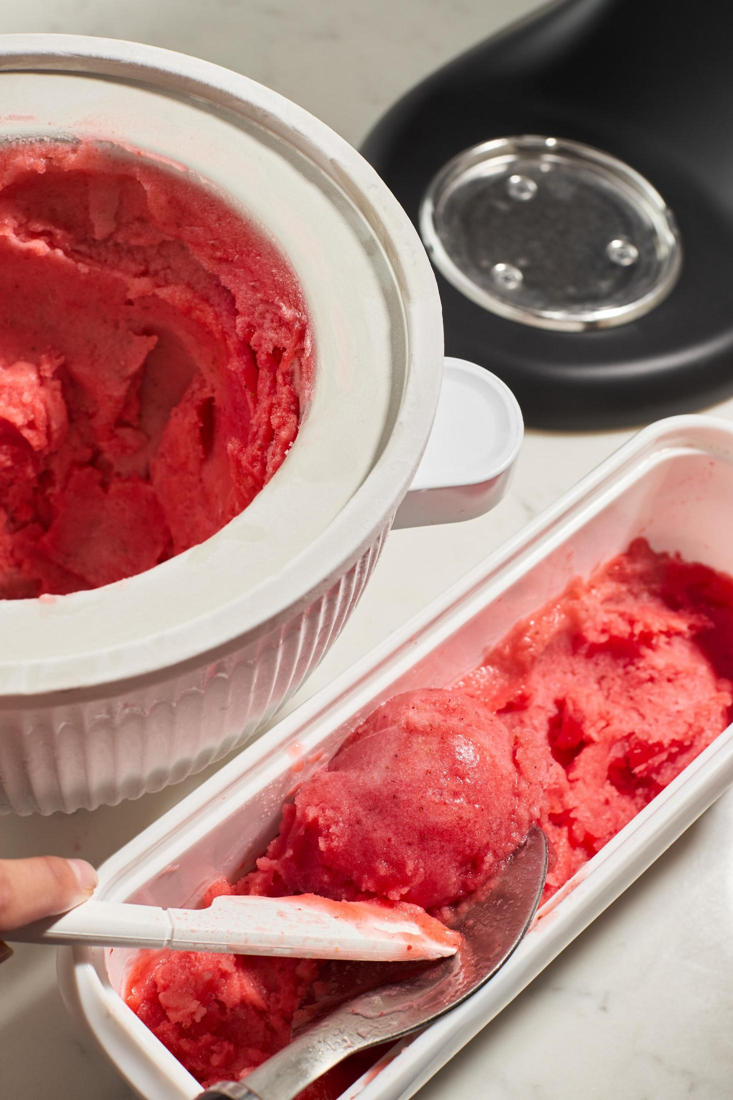 Rosa Eis in einem weißen Behälter, Löffel schöpft es, mit Eismaschine dahinter.