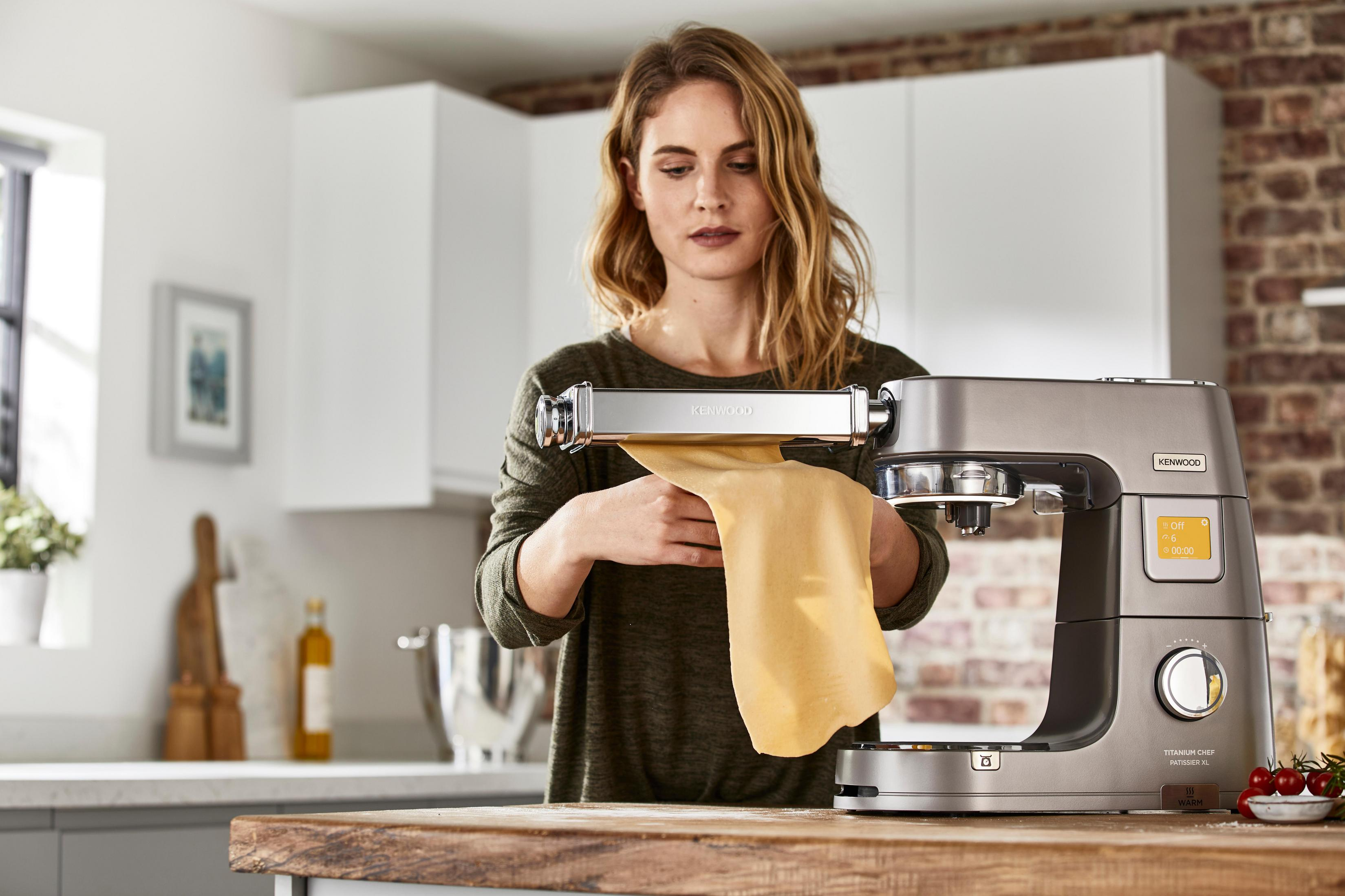 Kobieta używająca srebrnej maszyny kuchennej Kenwood do robienia makaronu w jasnej kuchni.