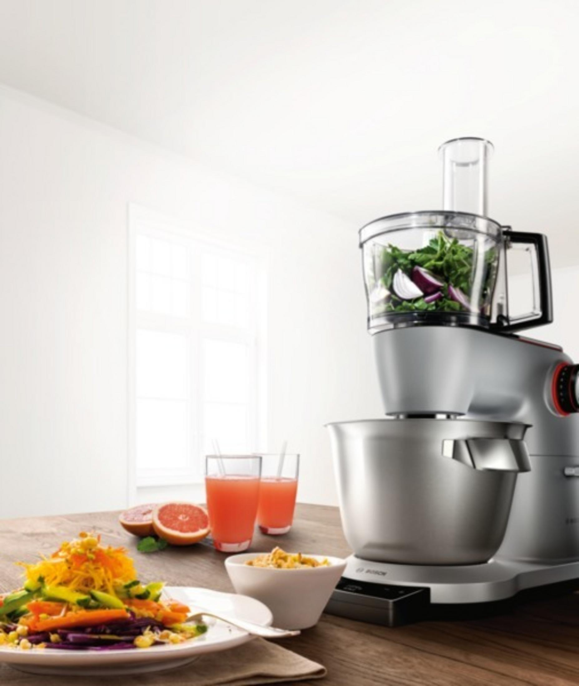 Eine Küchenmaschine mit Gemüse darin. Zwei Gläser Saft und ein Salat stehen auf dem Tisch.