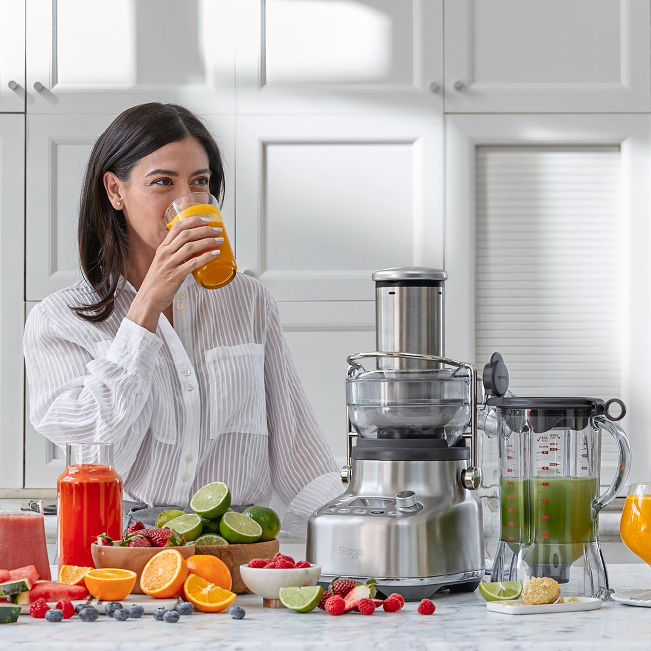 Kobieta pije sok. Wyciskarka, owoce i szklanki z sokiem na blacie. Białe szafki w tle.