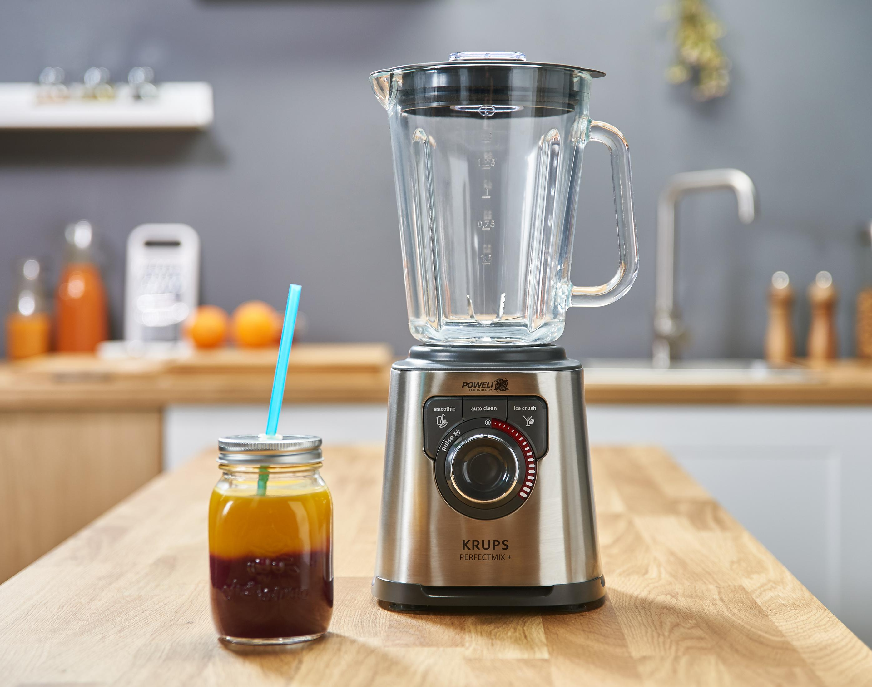 Mixer mit Glasbehälter und Smoothie-Glas mit Strohhalm auf einem Holztisch. Orangen im Hintergrund.