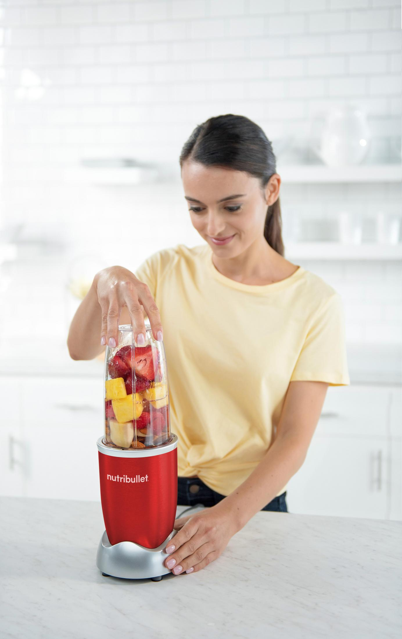 Mujer añadiendo fruta a una licuadora roja, preparando un batido en una cocina brillante.