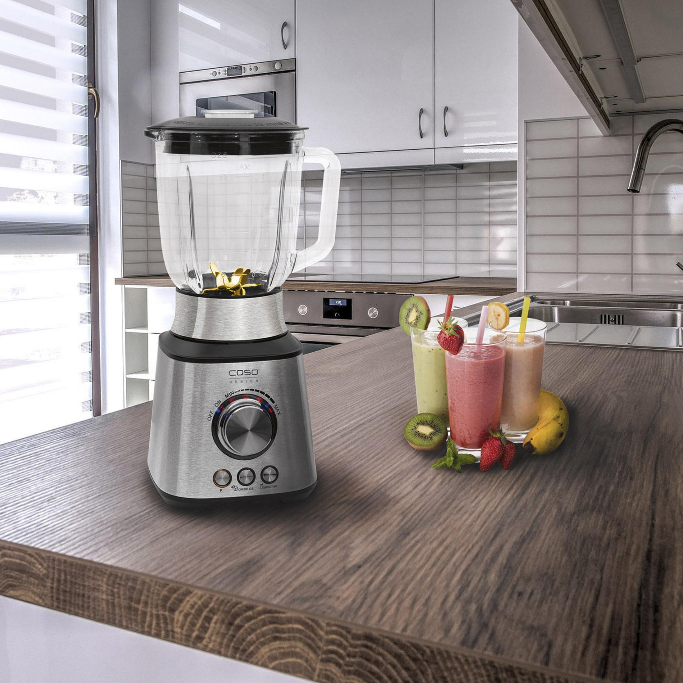 Ein Mixer auf einer hölzernen Theke mit Früchten und Smoothies daneben. Küchenschränke sind im Hintergrund.