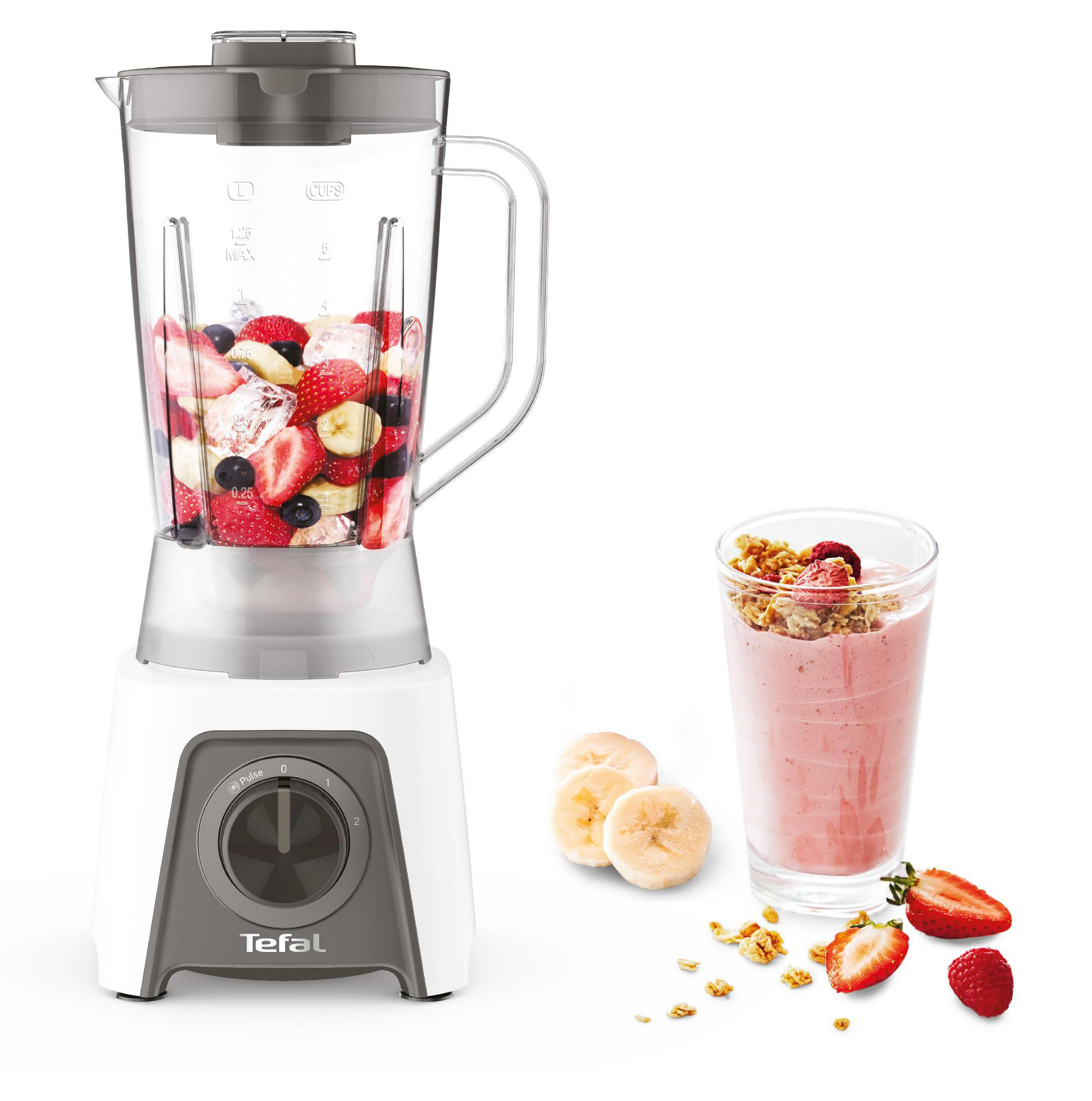 Una licuadora blanca y gris con frutas dentro. Un vaso de batido y plátanos al lado.