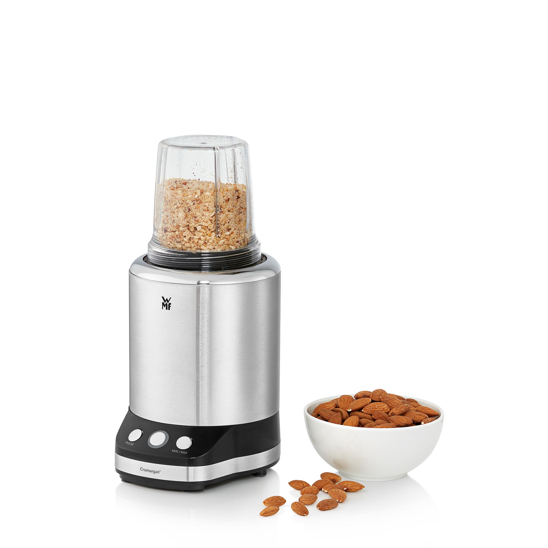 Srebrno-czarna elektryczna szlifierka do orzechów. Ma przezroczysty pojemnik wypełniony posiekanymi orzechami.