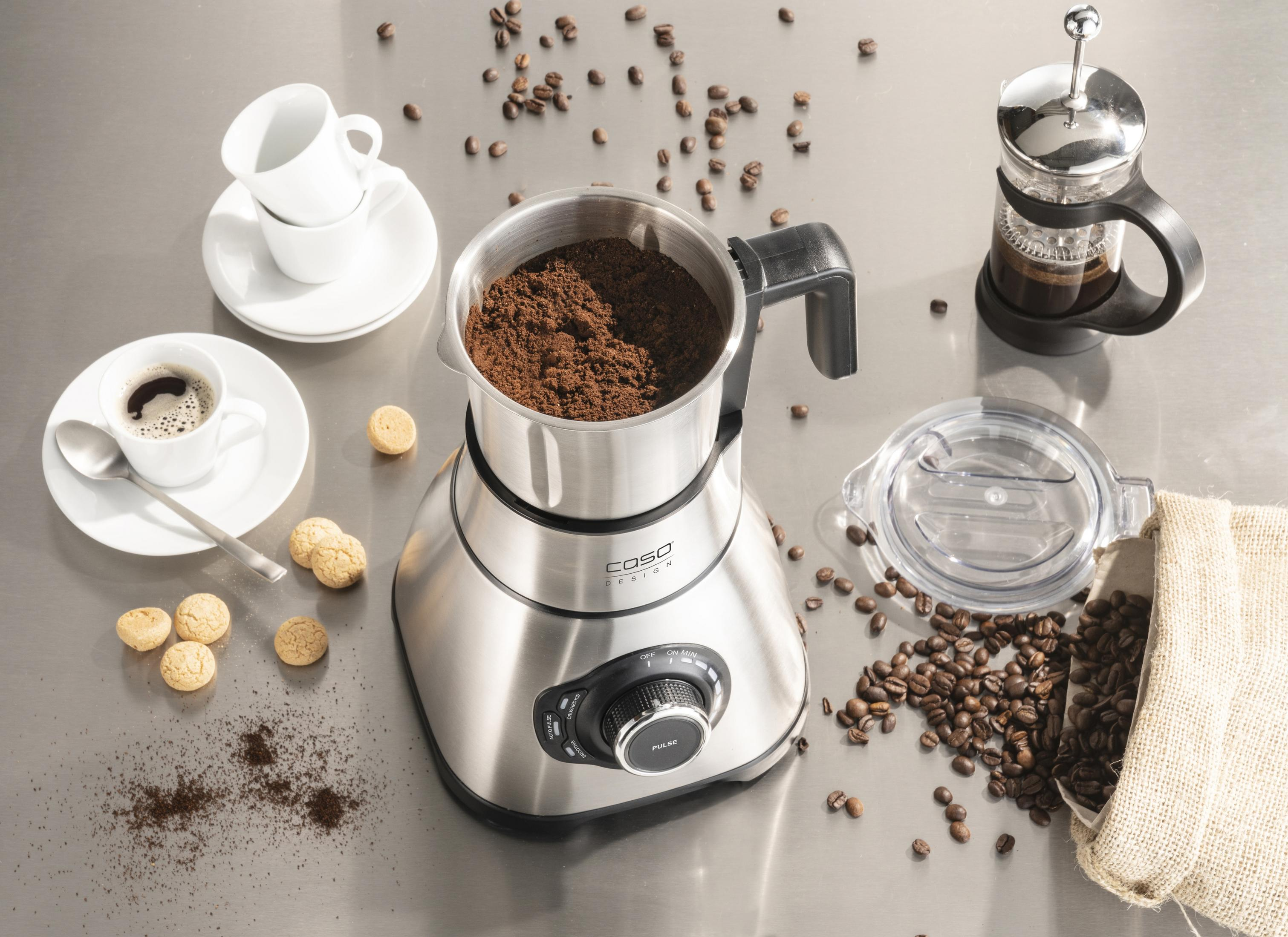 Kaffeemühle mit Kaffeebohnen, Tassen und Keksen auf metallischer Oberfläche.
