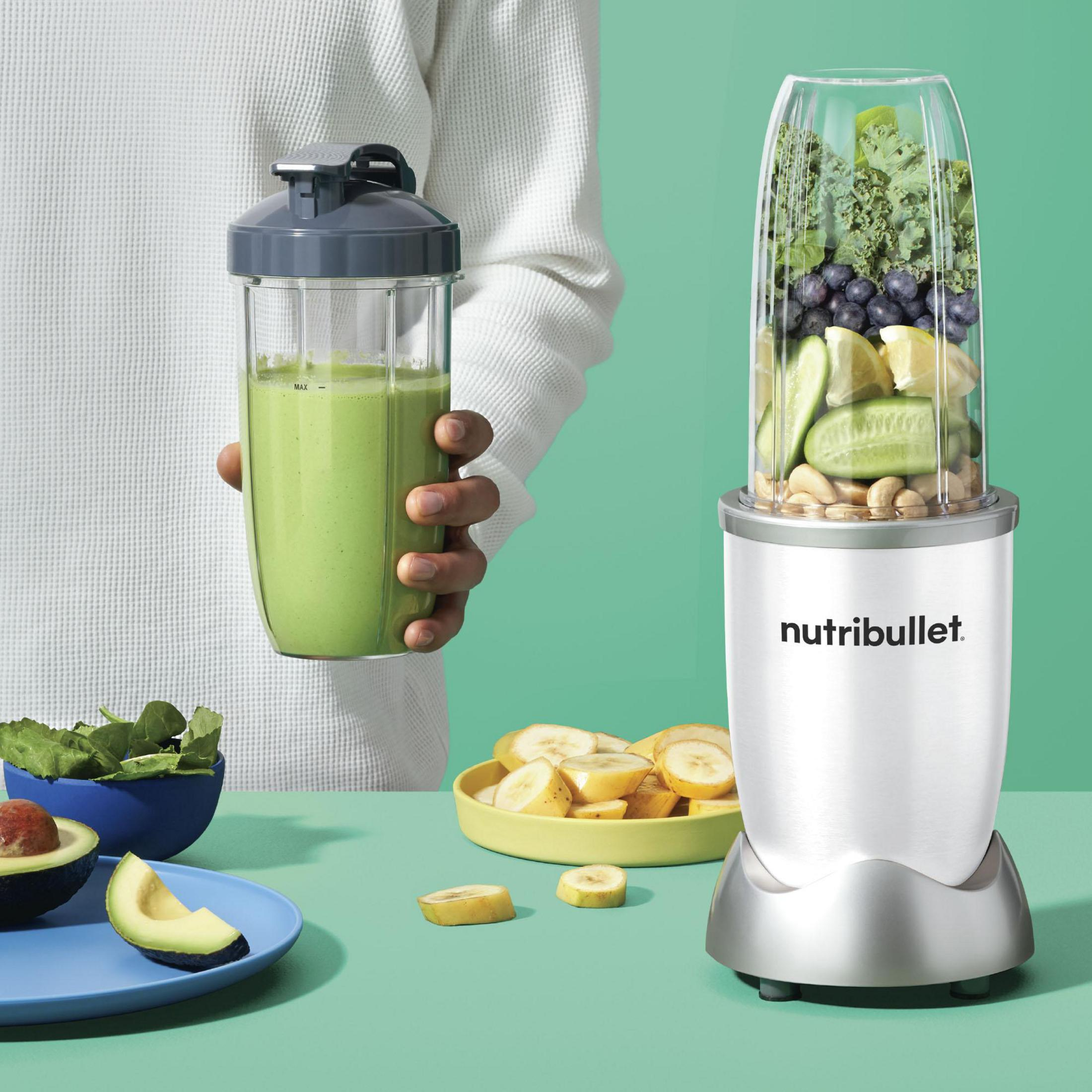Eine Person hält einen grünen Smoothie in einem Shaker. Ein Nutribullet und Zutaten daneben.