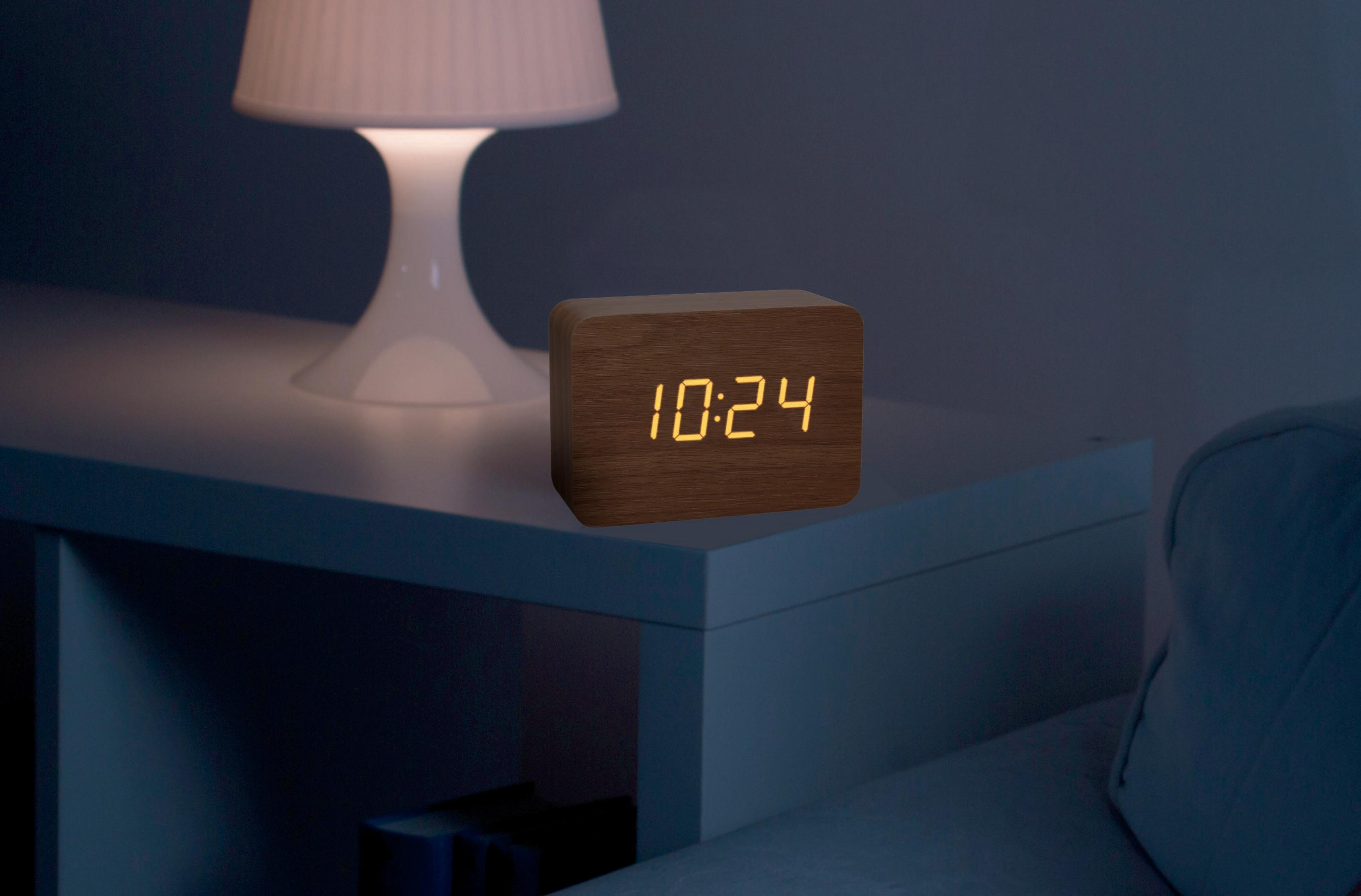 Wecker aus Holz mit Anzeige 10:24, auf einem weißen Tisch neben einer Lampe.