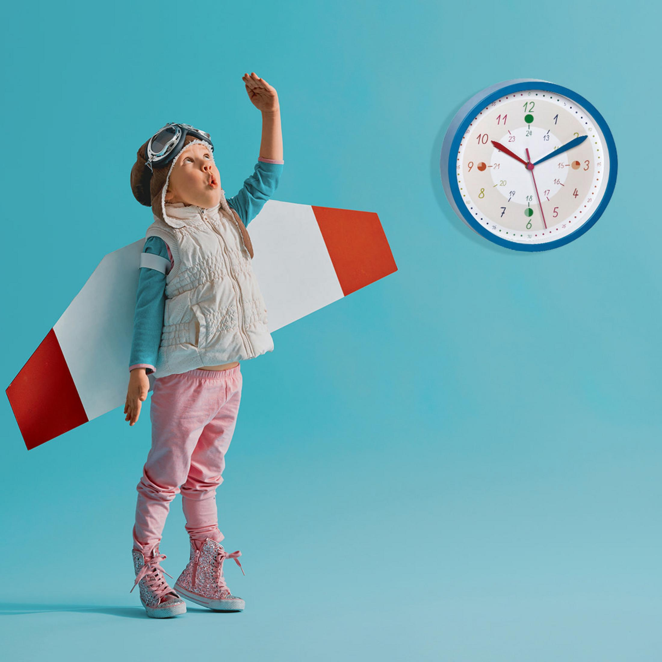 Ein Kind im Pilotenoutfit mit Flügeln blickt nach oben. Eine Uhr hängt an der blauen Wand.