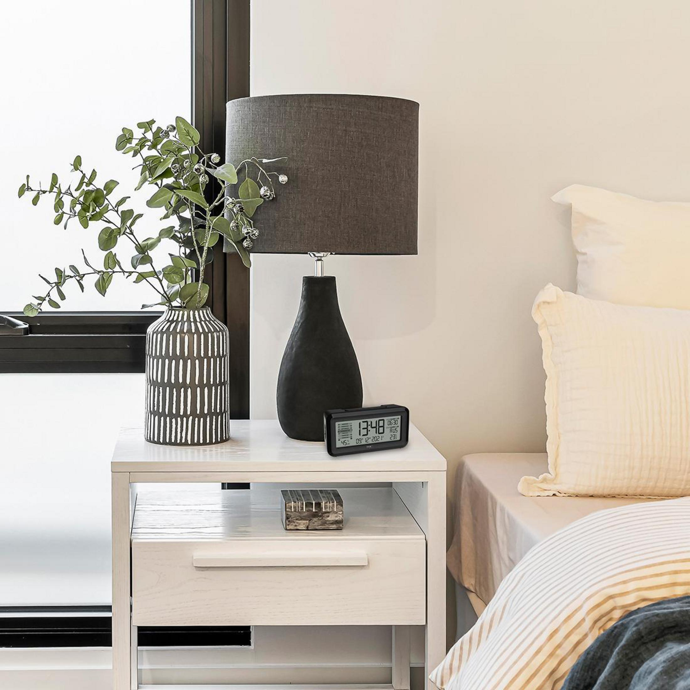 Nachttisch mit Lampe, Blumenvase und Digitaluhr. Bett im Hintergrund.