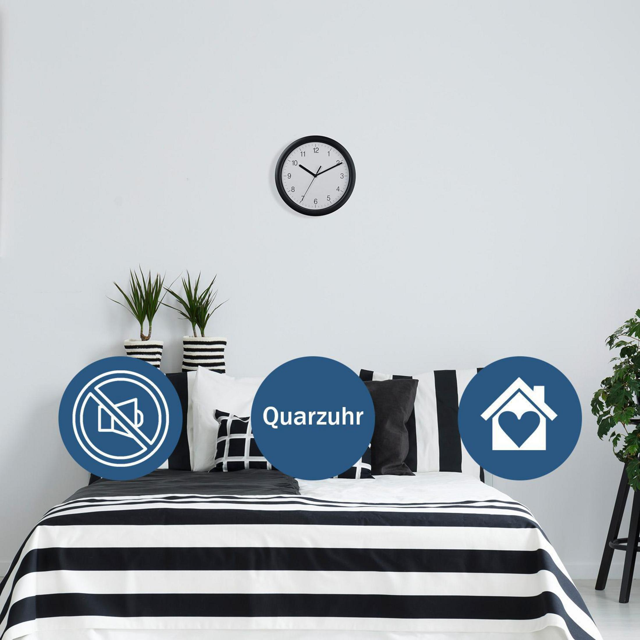 Eine Uhr an einer weißen Wand über einem Bett. Es gibt Symbole und Pflanzen.