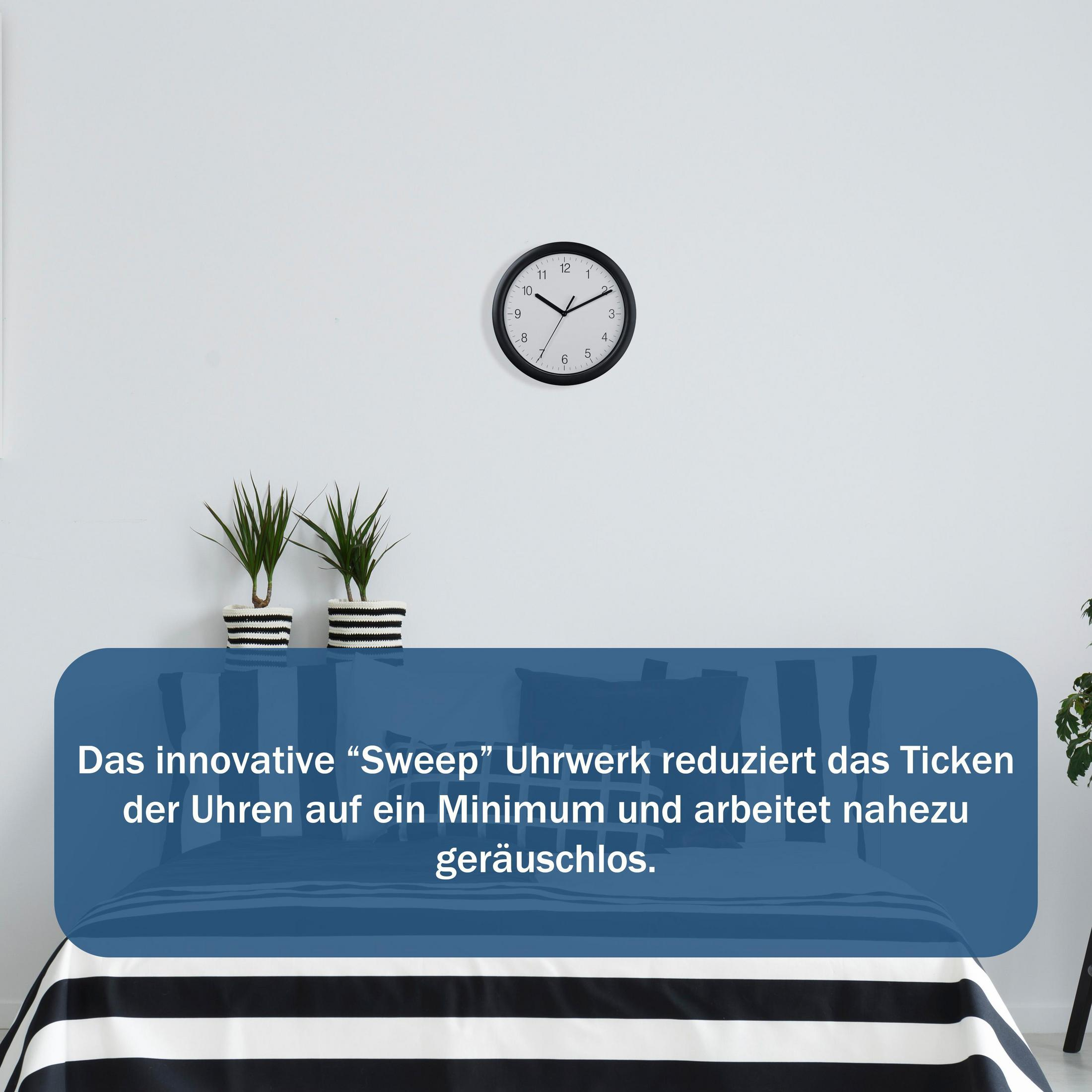 Eine schwarze Uhr an einer weißen Wand über einem Bett mit gestreiften Laken und zwei Topfpflanzen.