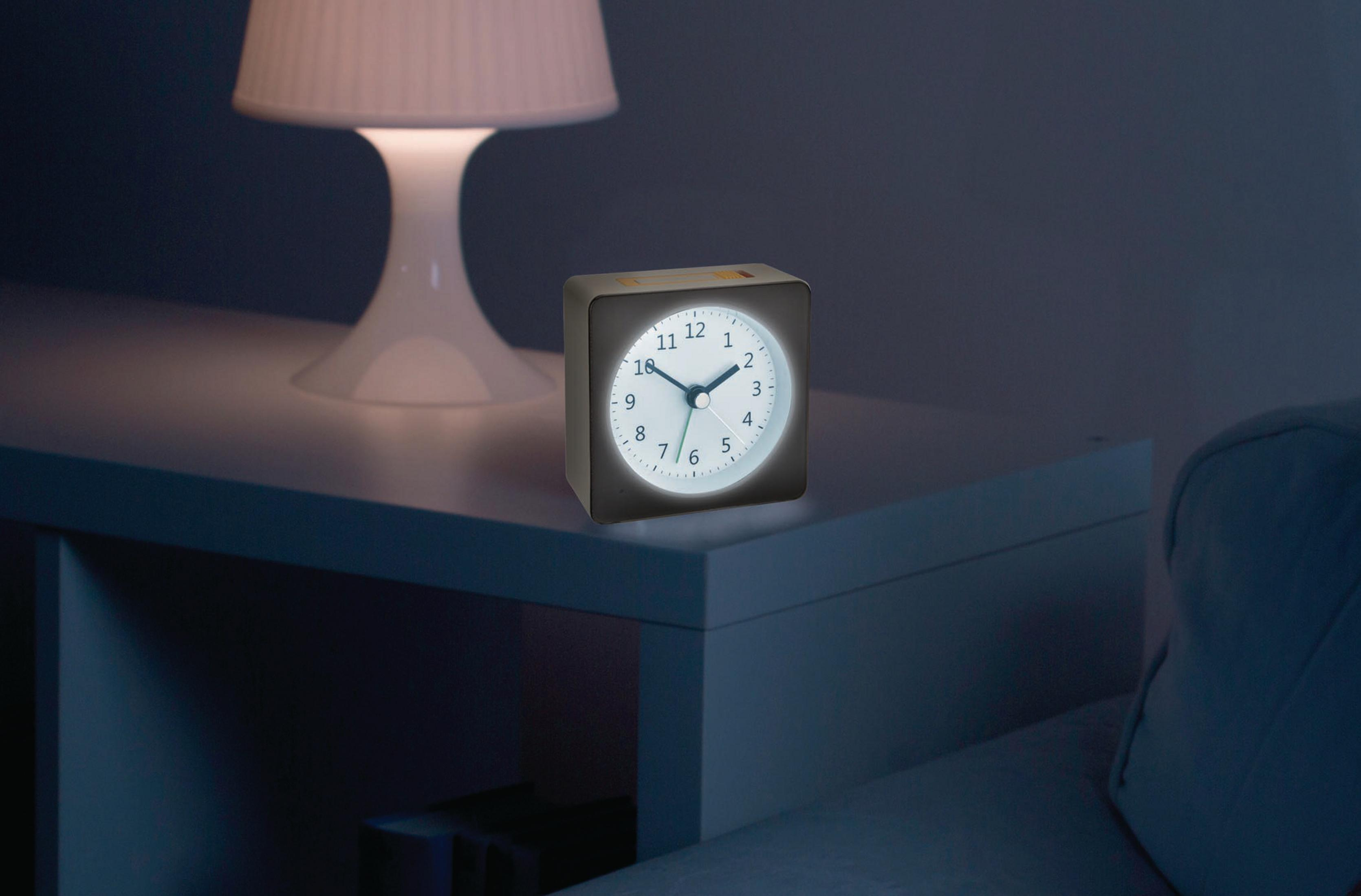 Ein kleiner, beleuchteter Wecker steht auf einem weißen Nachttisch neben einer weißen Lampe und einem Teil eines Sofas.
