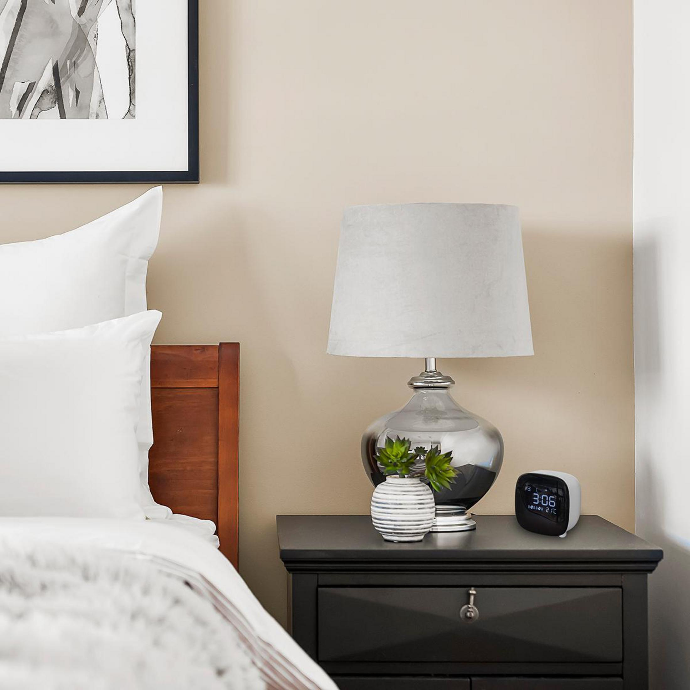 Scena della camera da letto: lampada, comodino con orologio, cornice, letto. Muro beige.