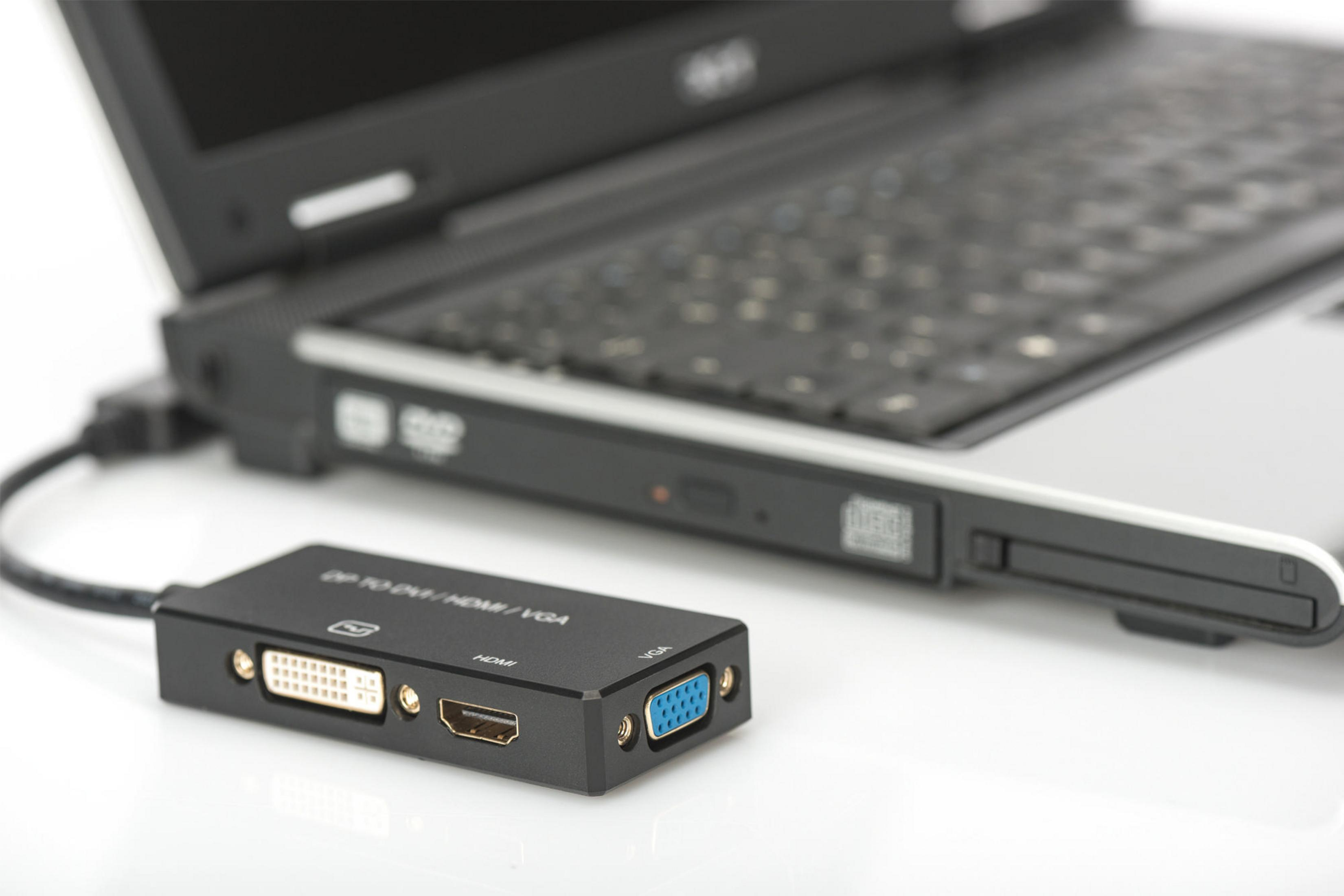 Schwarzer Adapter mit HDMI-, VGA- und DVI-Anschlüssen, neben einem Laptop. Weißer Hintergrund.