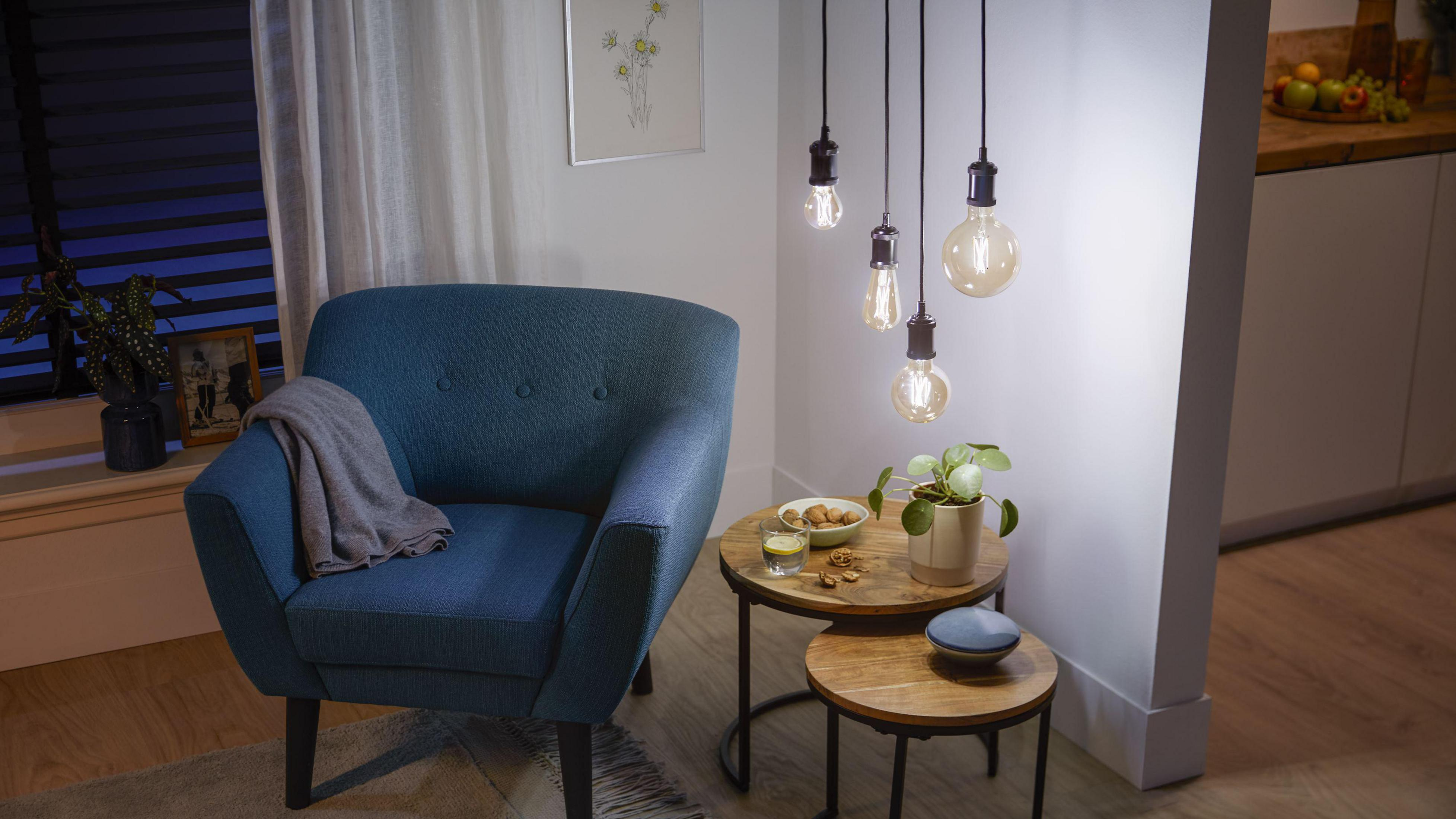 Niebieski fotel stoi obok drewnianych stolików z wiszącymi żarówkami. Obecne są rośliny i świece.